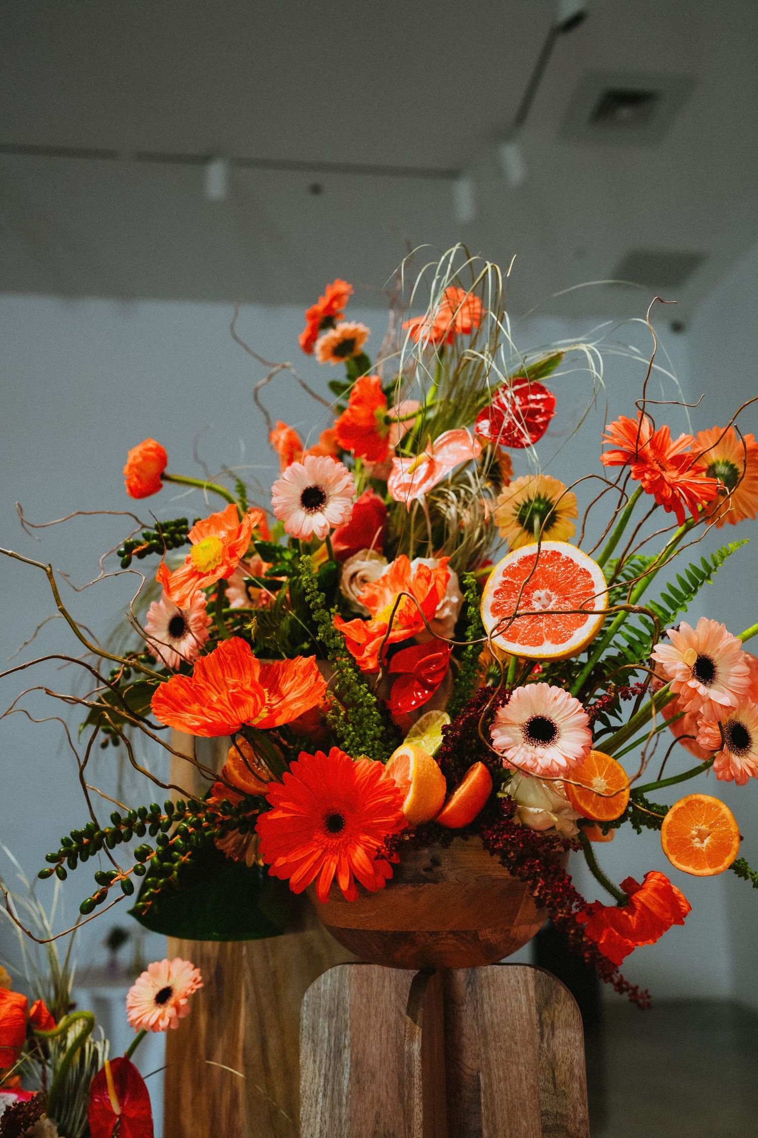 Vibrant orange floral arrangement with gerbera daisies, anthuriums, and citrus slices in a rustic wooden vase.