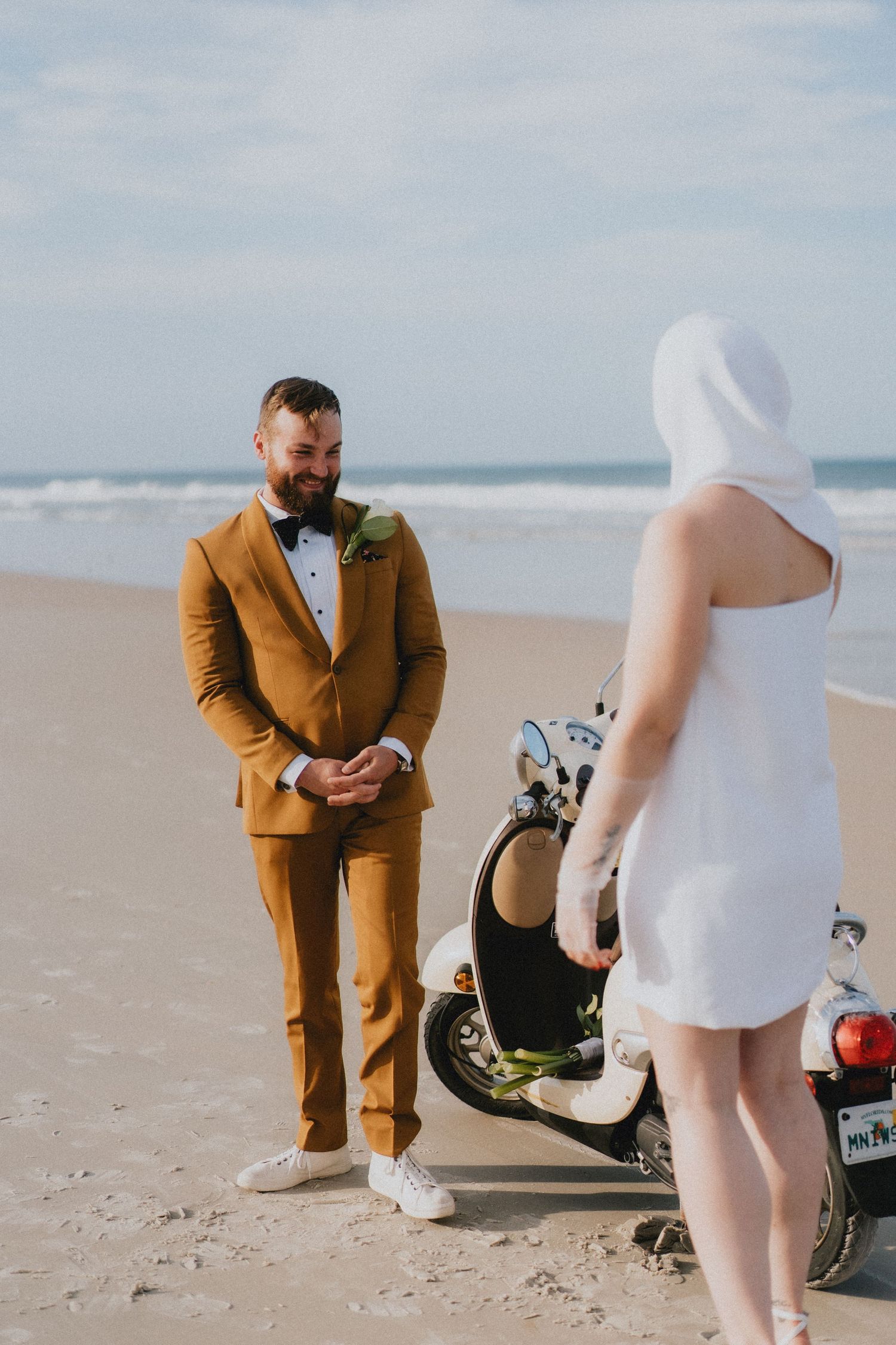 Groom in mustard suit smiling at bride in white mini dress on a sandy beach with a scooter.
