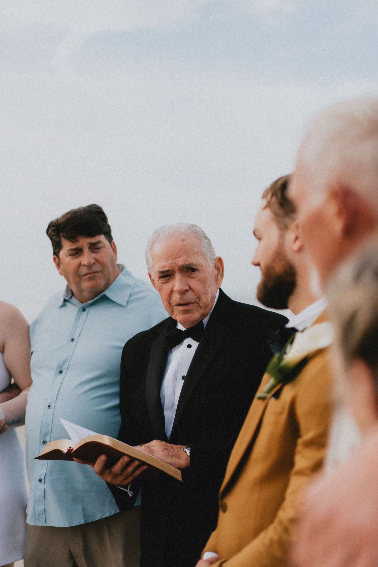 Elderly officiant in tuxedo reads from book during outdoor beach wedding ceremony.