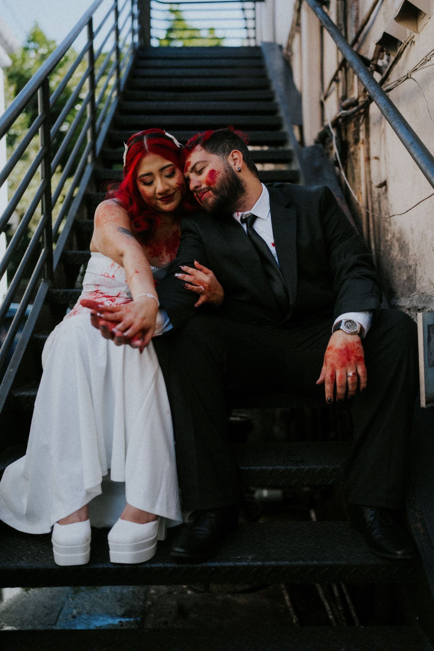 Zombie-themed wedding couple in bloody Halloween costumes sitting on metal stairs embracing romantically.