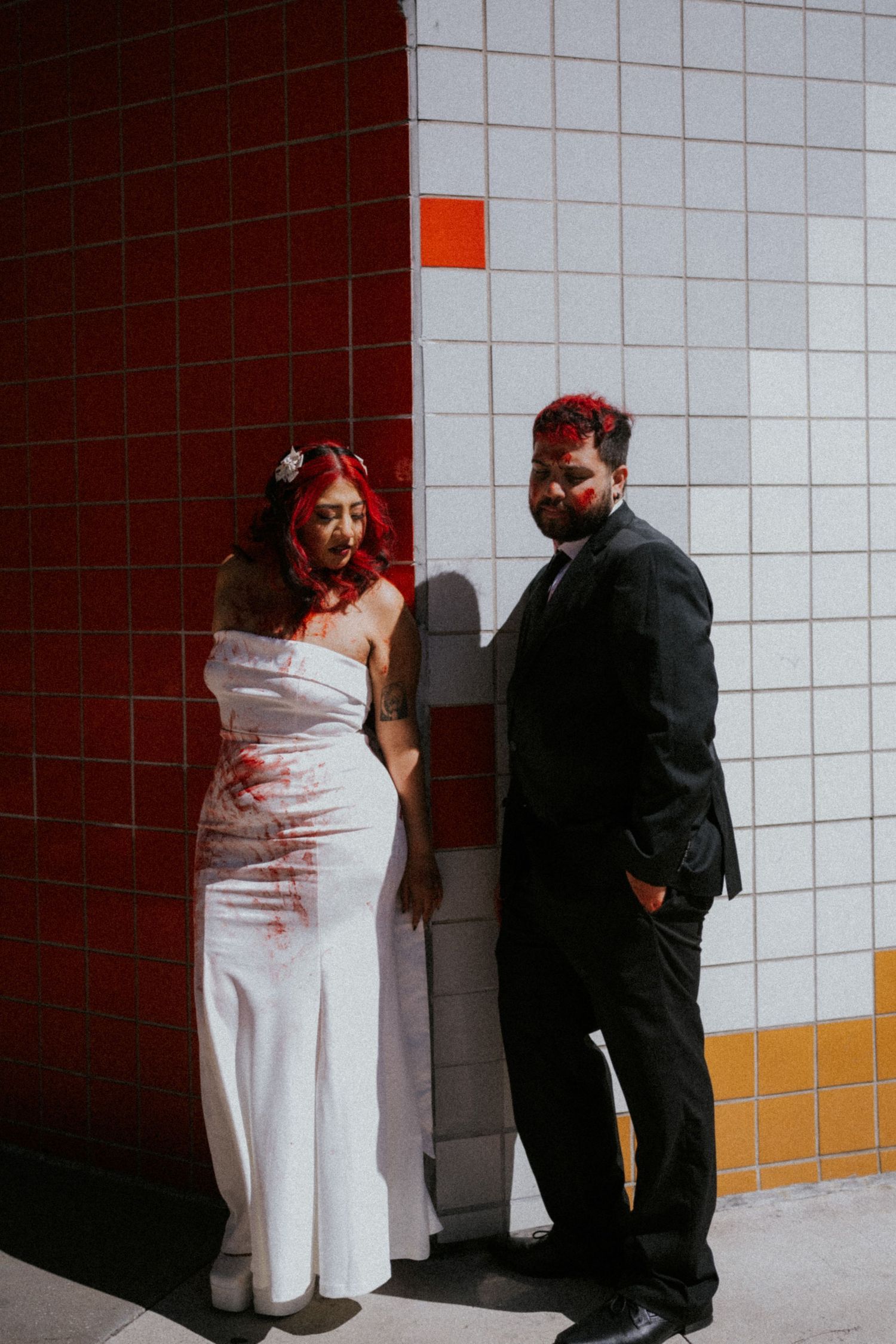 Bride in white gown and groom in black suit posed against colorful tiled wall with Halloween zombie makeup.