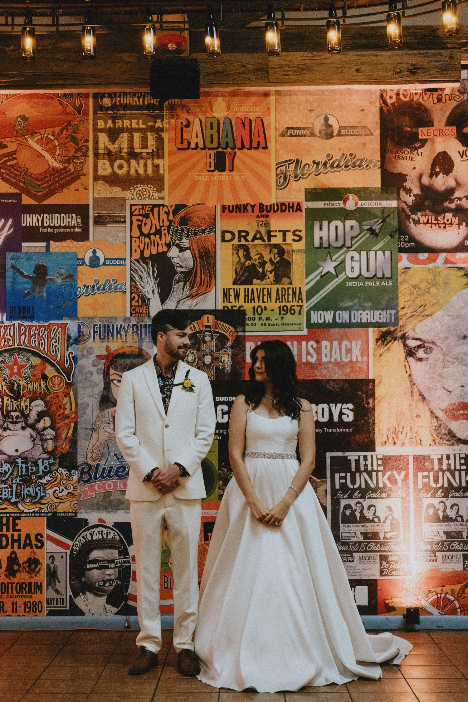 Bride and groom pose in white wedding attire before a colorful vintage concert poster wall with string lights above.