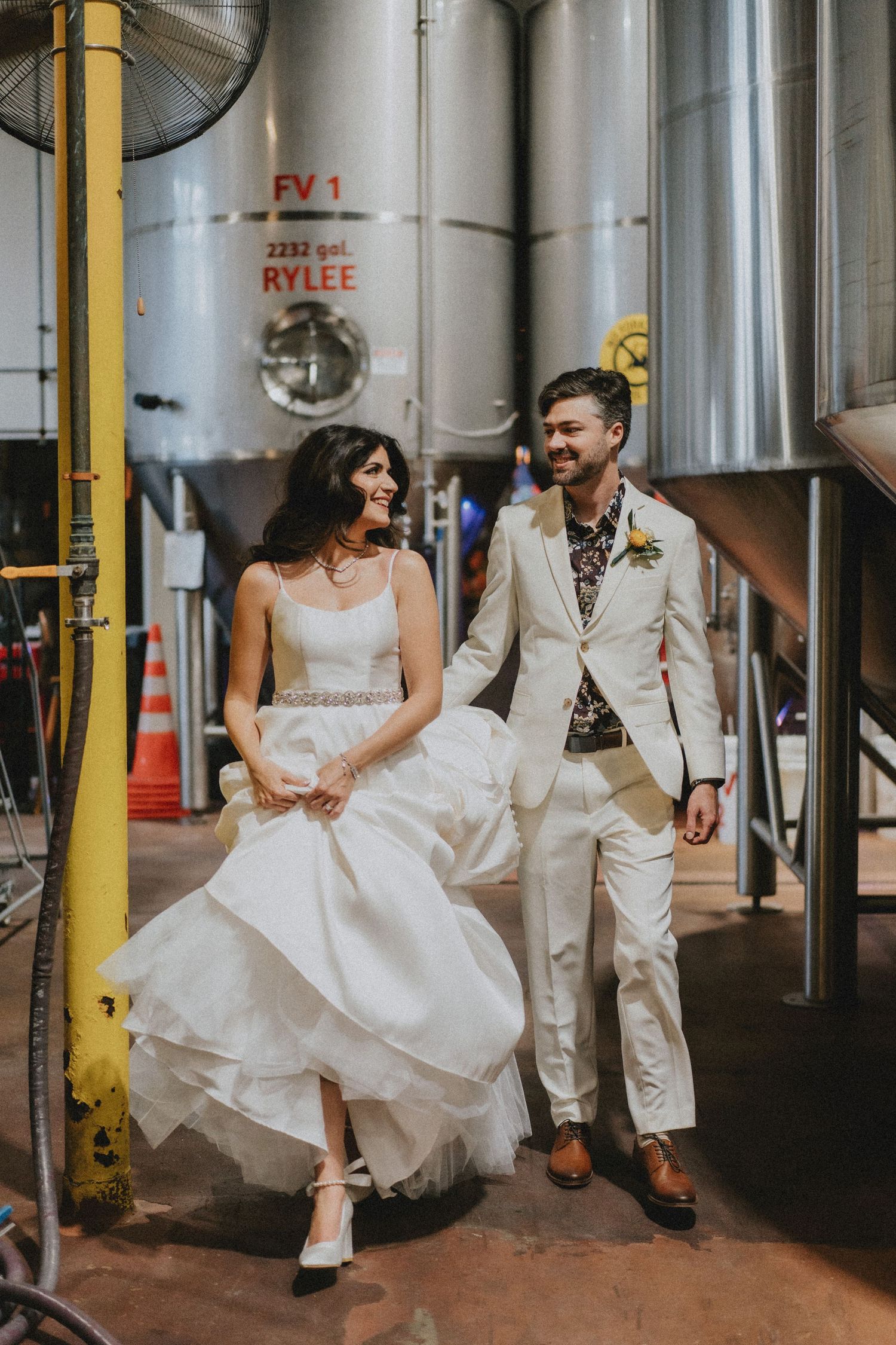 Bride and groom walking together laughing in a brewery wedding venue with large fermentation tanks.
