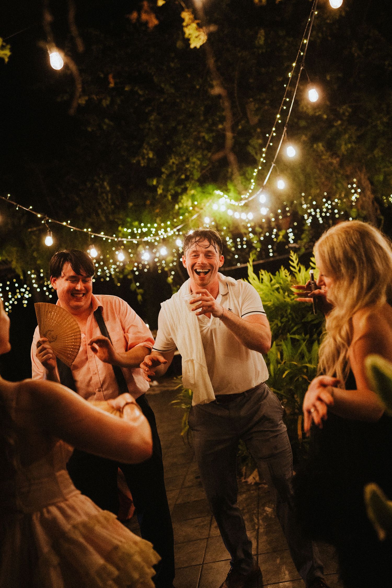 Guests laugh and dance outdoors at a nighttime wedding reception under warm string lights surrounded by lush greenery.