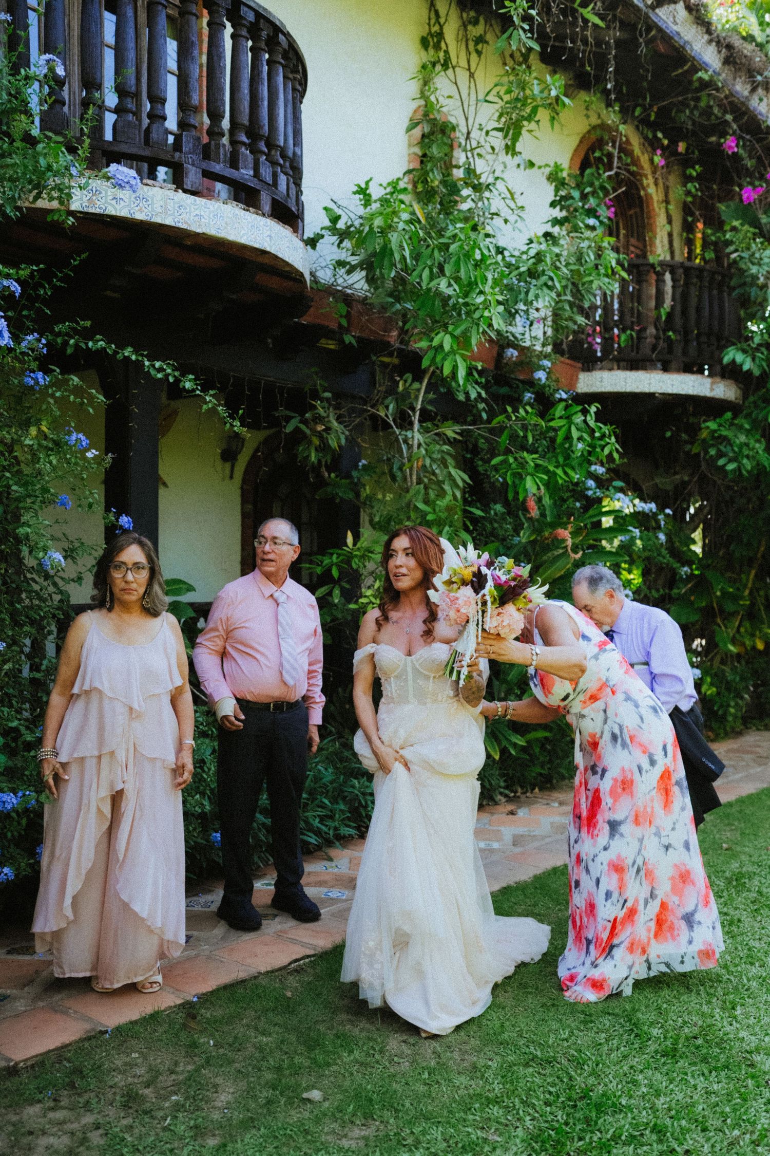 Bride in white gown receives bouquet from guest in floral dress at outdoor garden wedding venue with lush greenery.