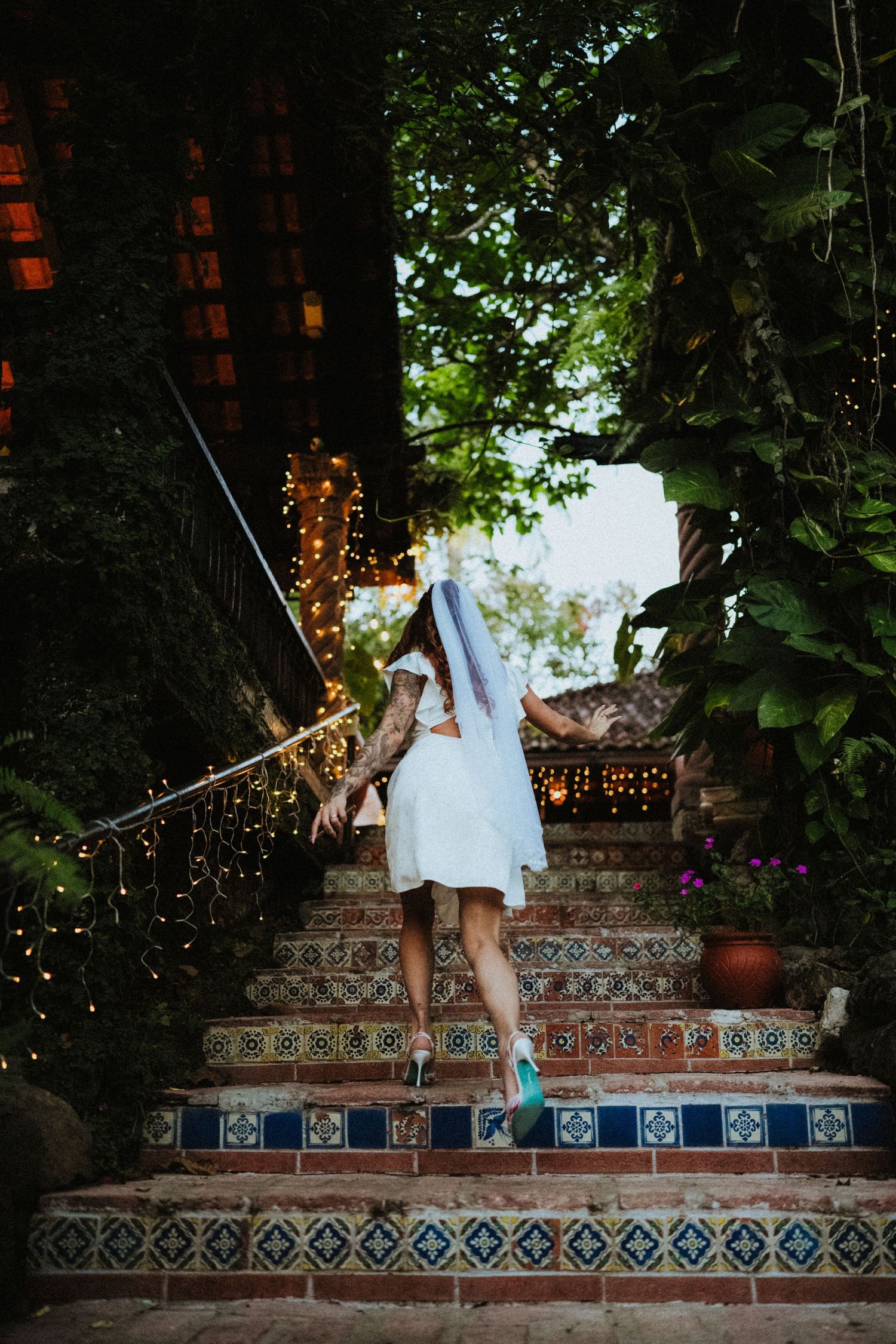Bride in short white dress and veil ascends decorative tiled stairs lined with fairy lights in lush garden.