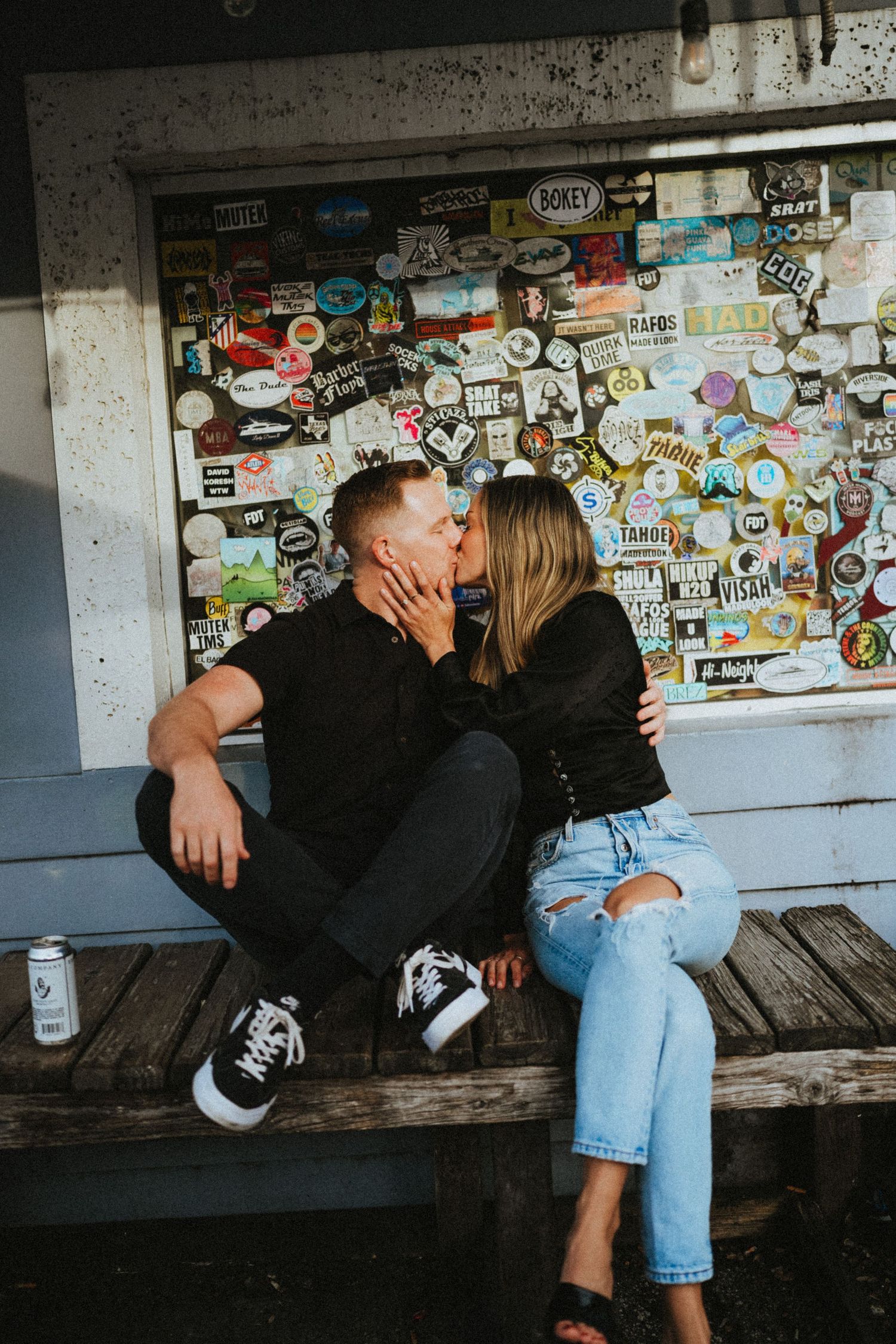 Couple kissing while sitting on a wooden bench in front of a colorful sticker-covered wall outdoors.