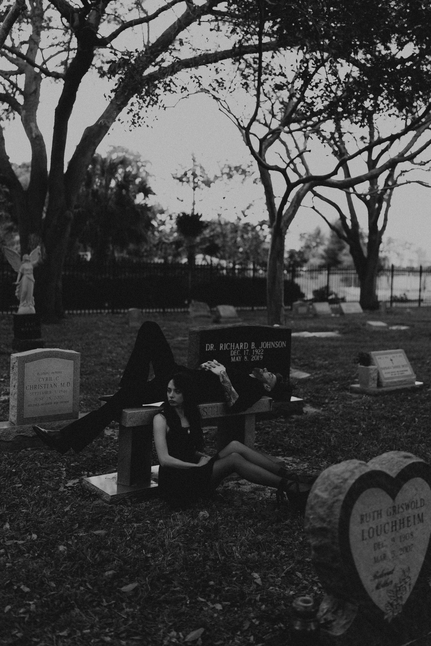 Black and white photo of a person resting among gravestones in a cemetery with large trees.