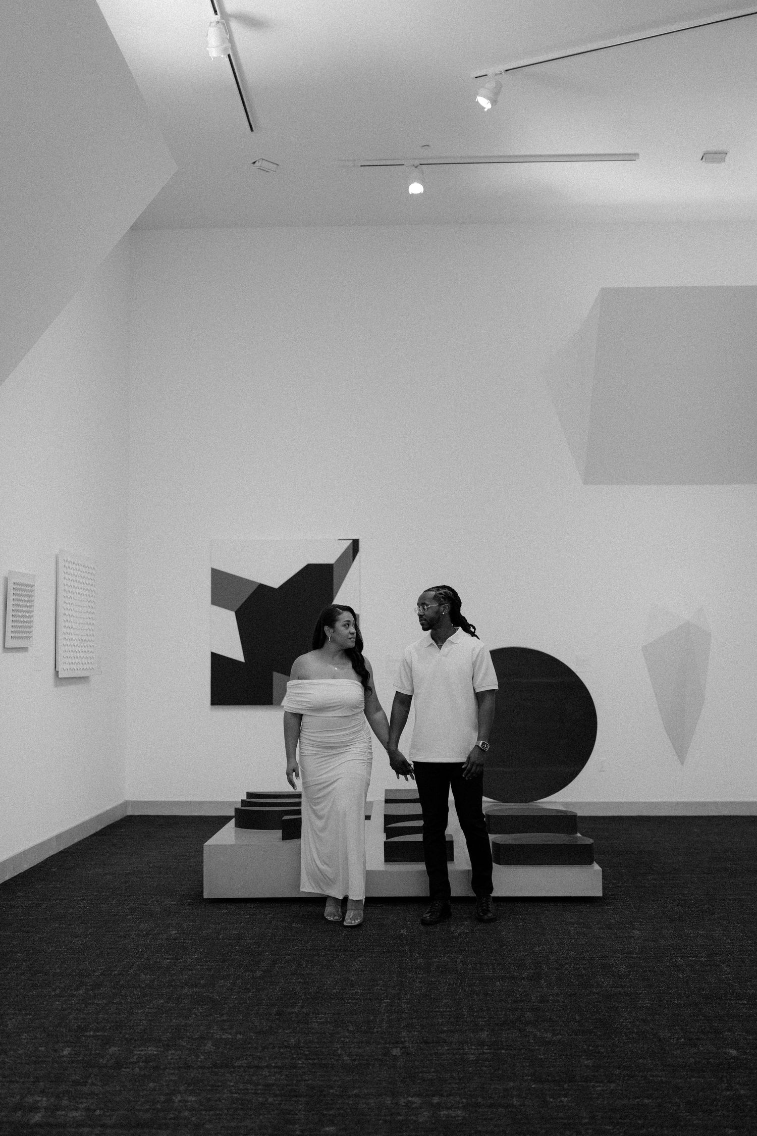 Black and white photo of a couple holding hands in a modern art gallery with abstract sculptures and artwork.