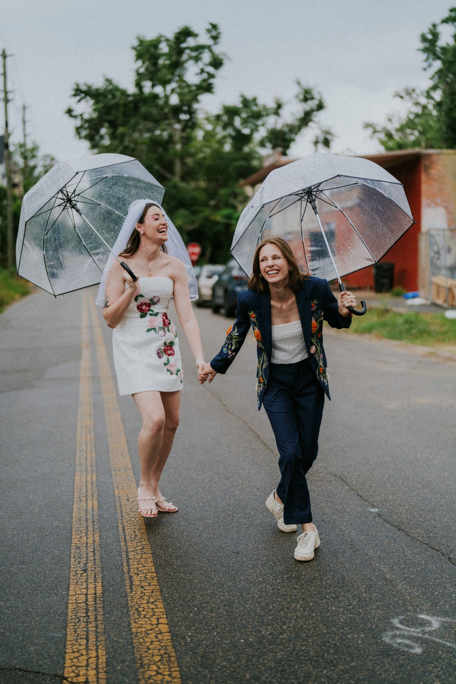 Two brides holding clear umbrellas walk joyfully hand-in-hand down a road on a rainy wedding day.