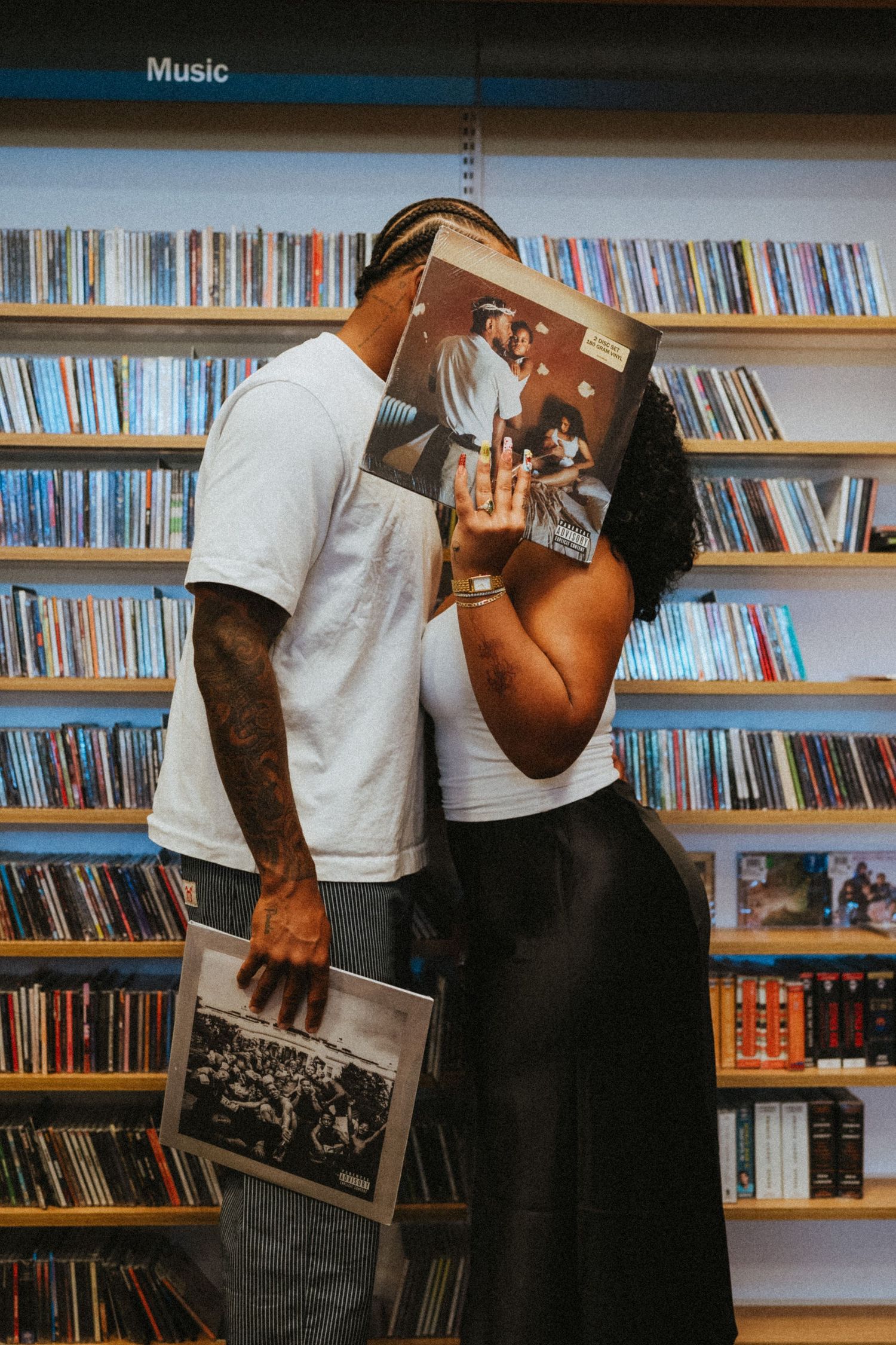 A couple kisses behind a vinyl record in a music store with shelves of albums in the background.