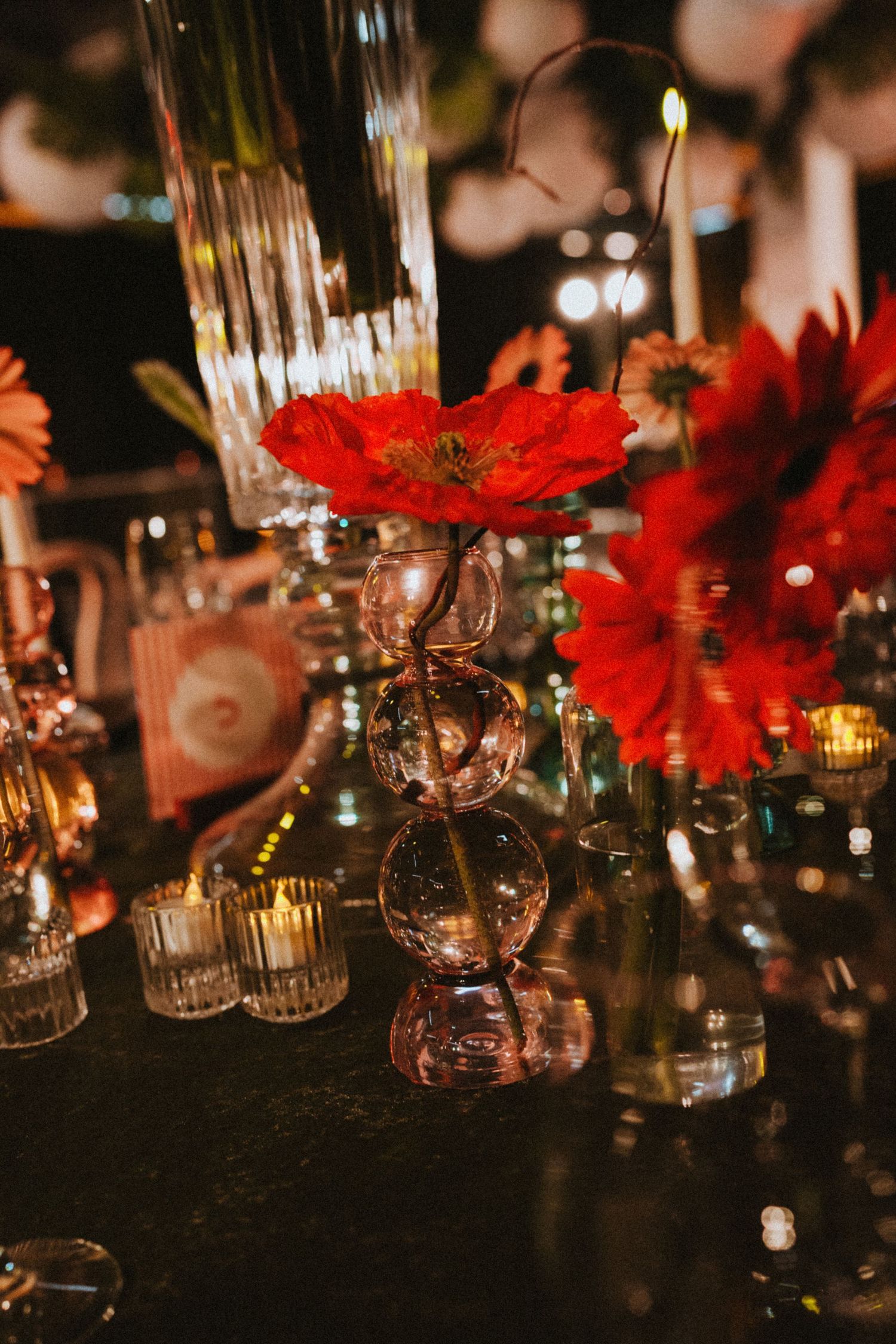 Red poppy and gerbera daisy centerpieces in glass bubble vases on dark table with candles.