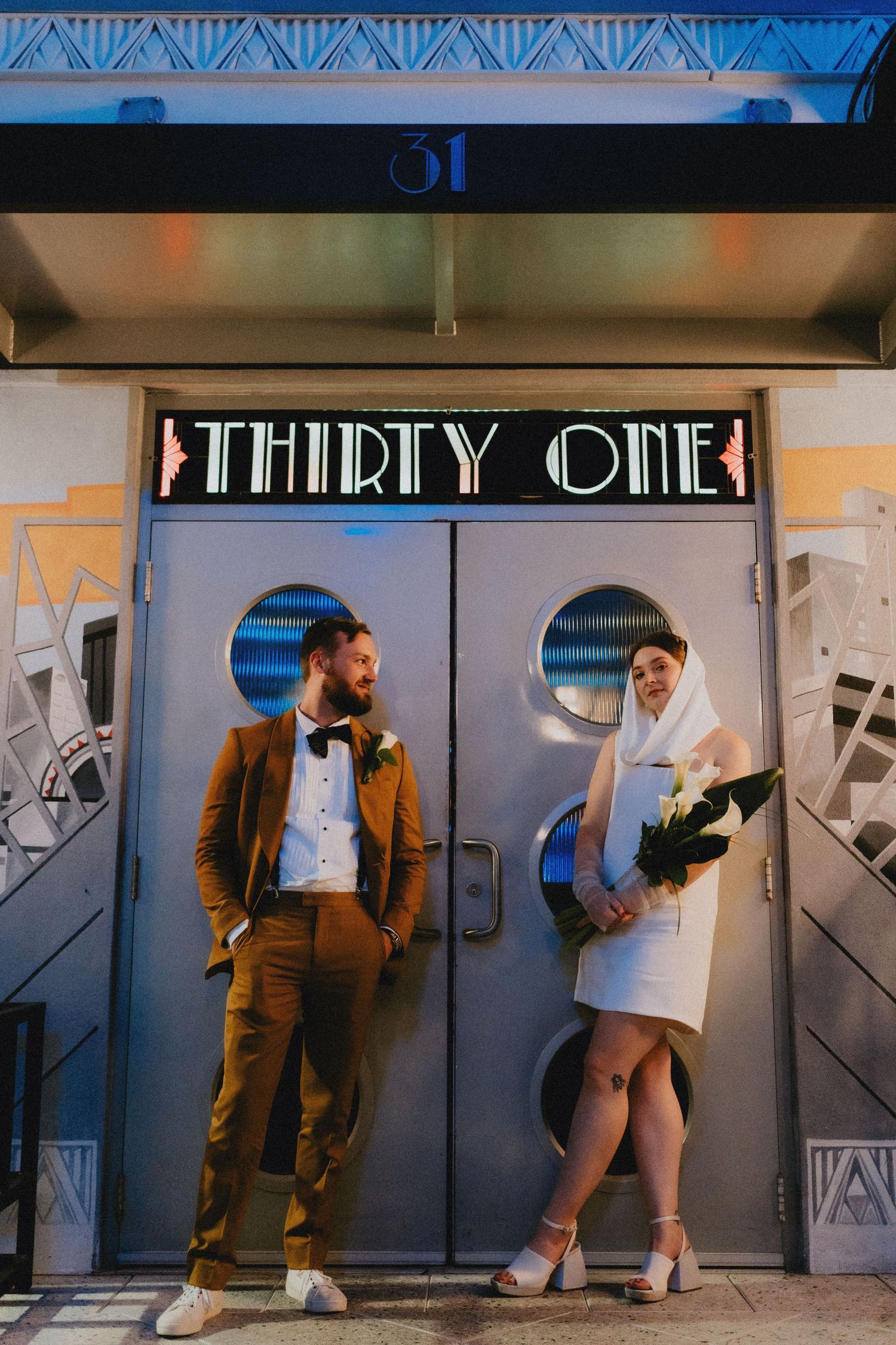Wedding couple poses outside Thirty One venue, groom in brown suit, bride in white mini dress.