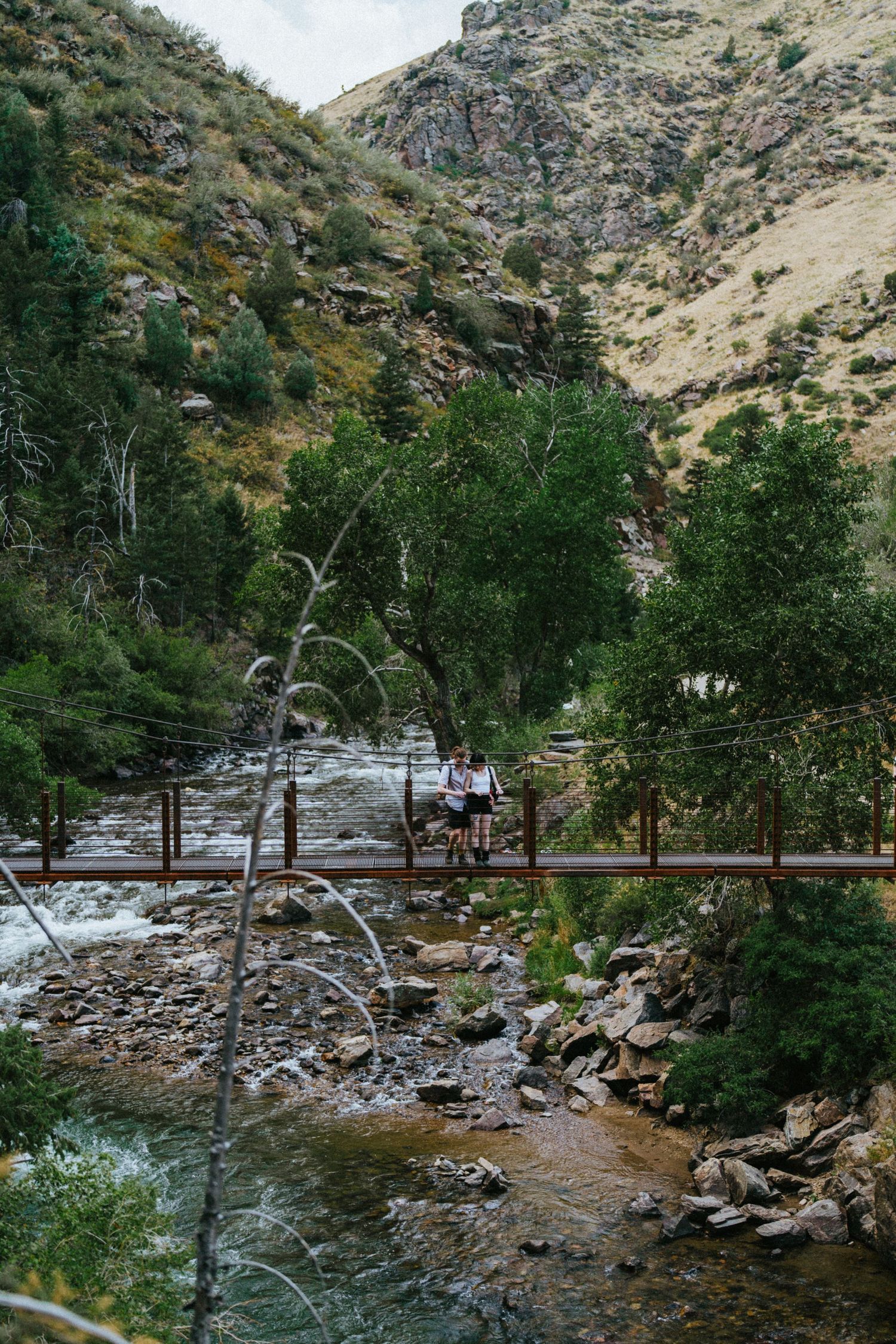 Couple standing on a wooden footbridge over a rocky mountain stream surrounded by lush green trees.