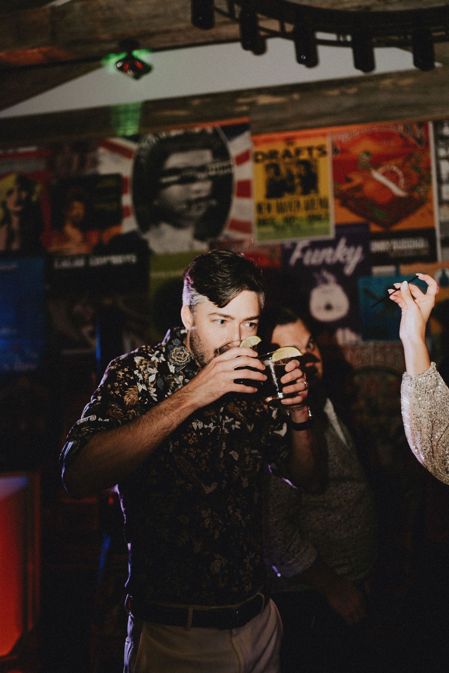 Man in floral shirt drinking a dark beer at a bar with vintage posters on the wall.