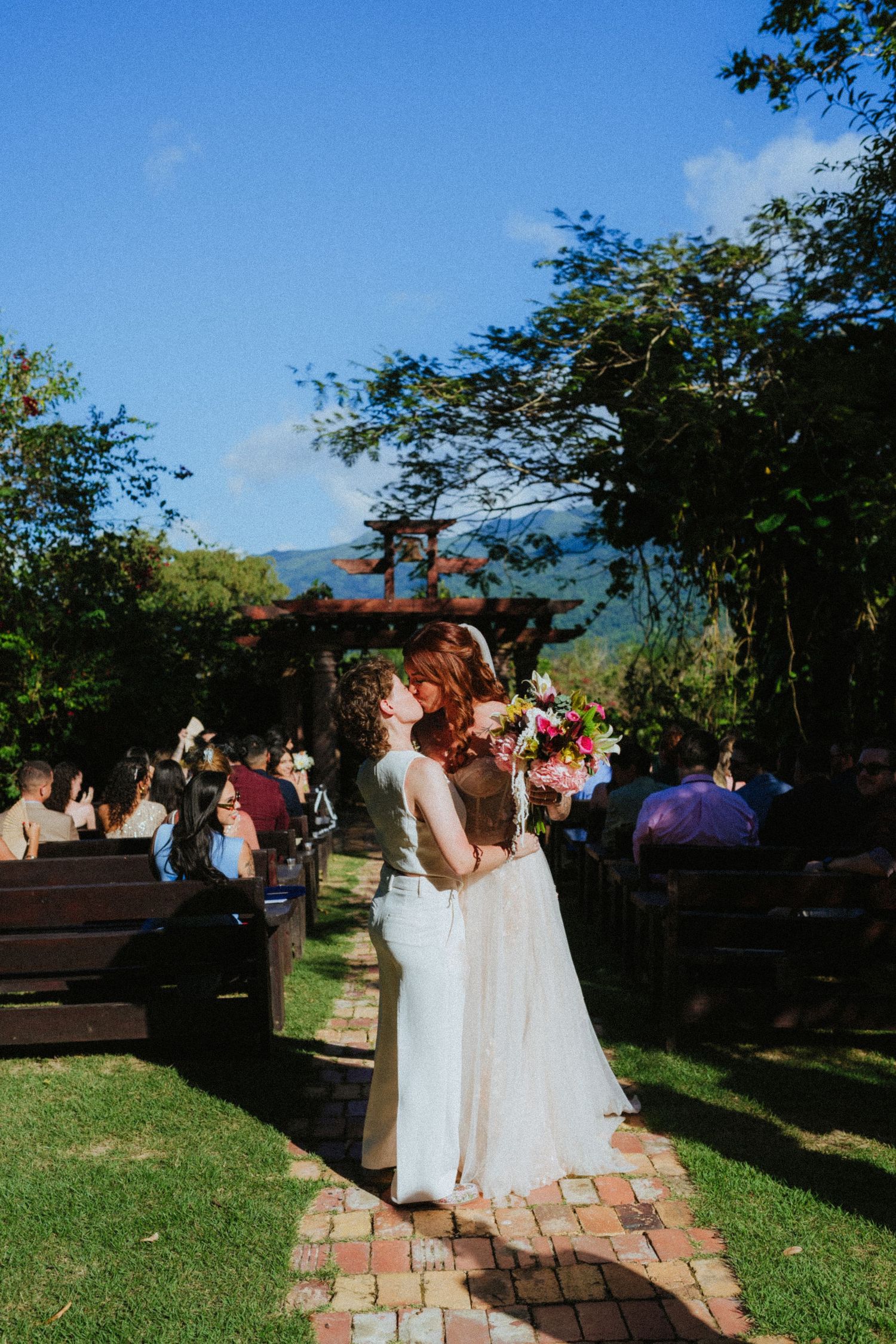 Two brides share a kiss at outdoor wedding ceremony, holding colorful bouquet on brick path surrounded by lush greenery.