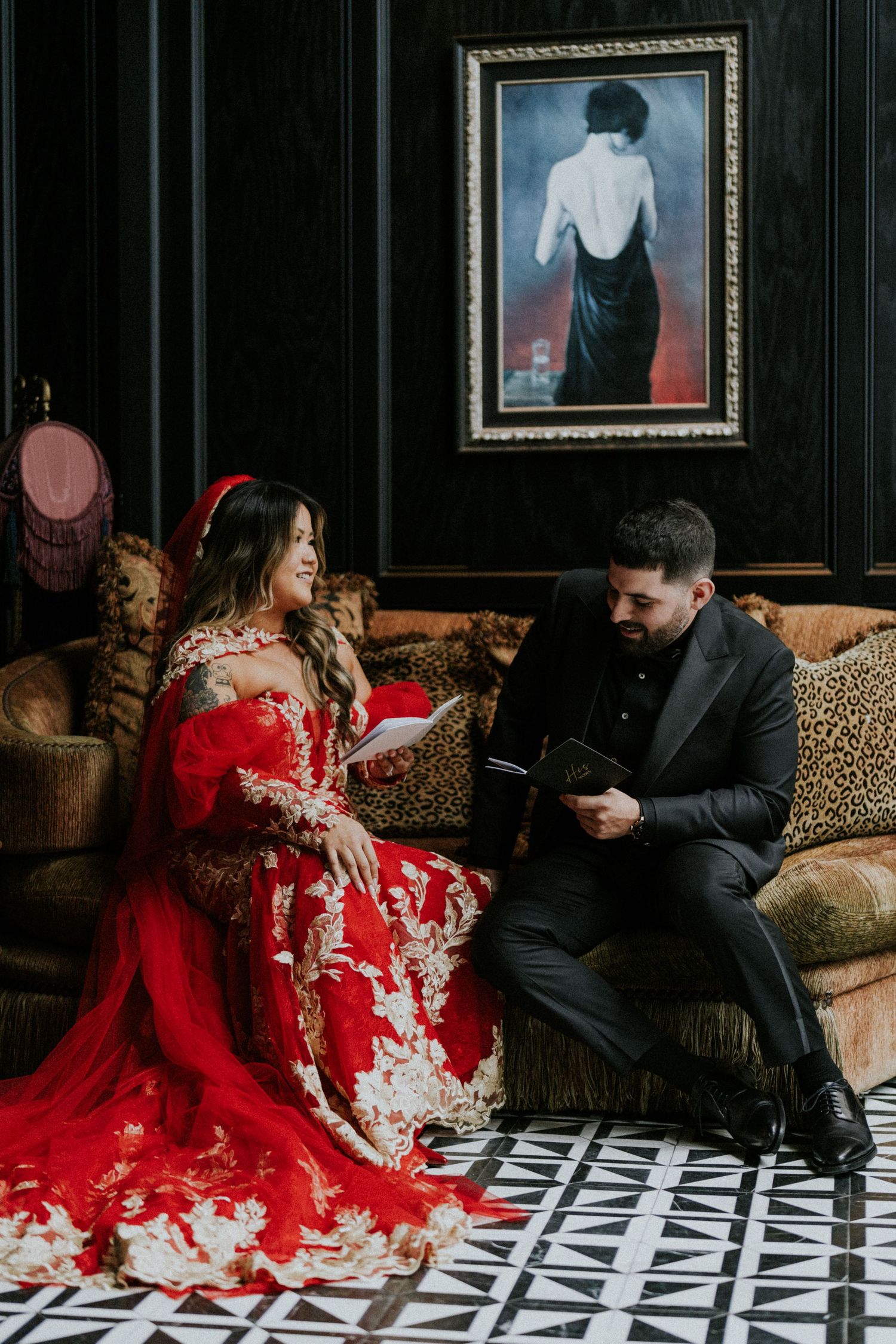 Couple in elegant attire sitting on ornate sofa, woman in red lace gown, man in black suit, reading vows in dark luxe room.