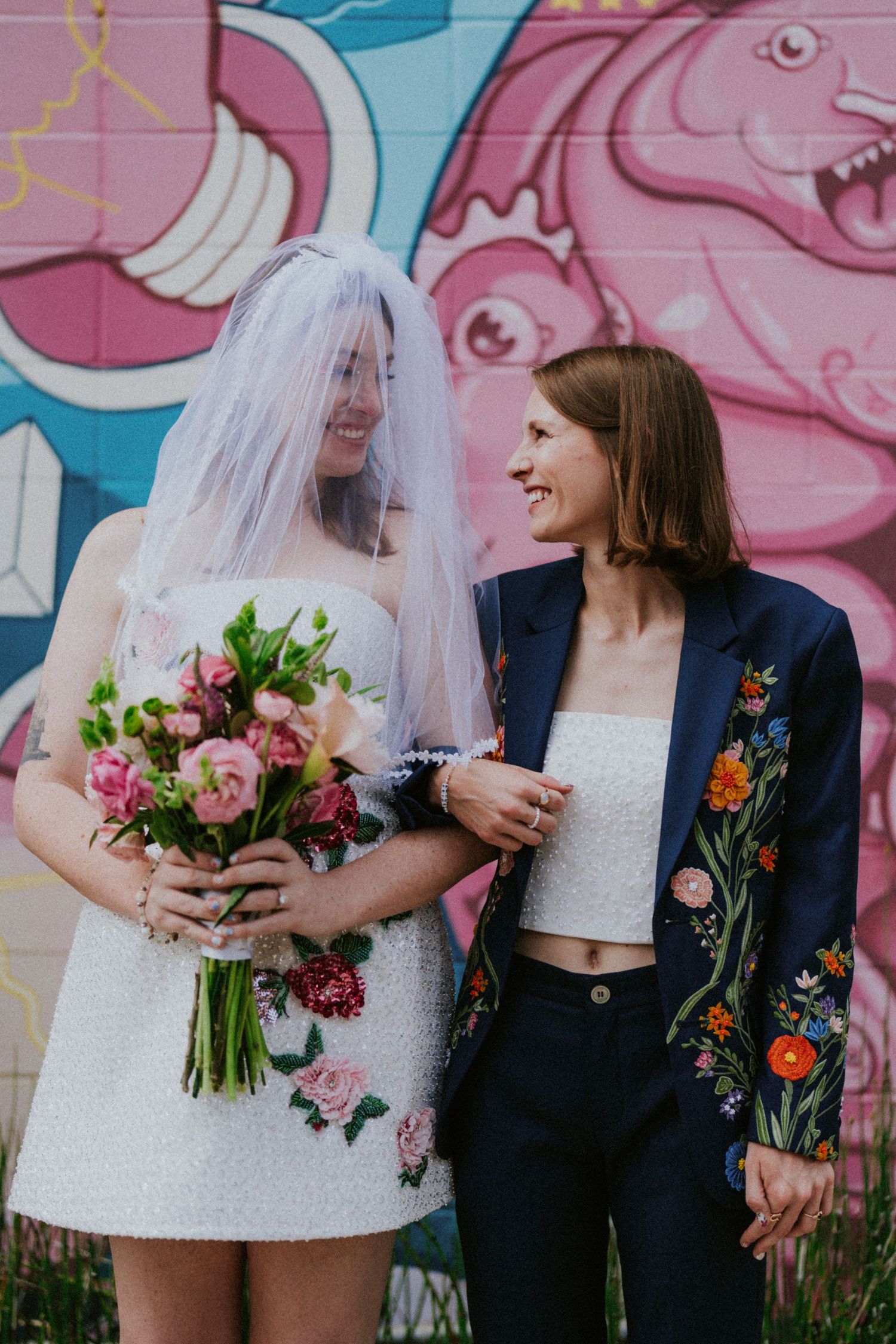 Two brides smile at each other, one in a floral mini dress with veil holding a pink bouquet, the other in an embroidered blazer.
