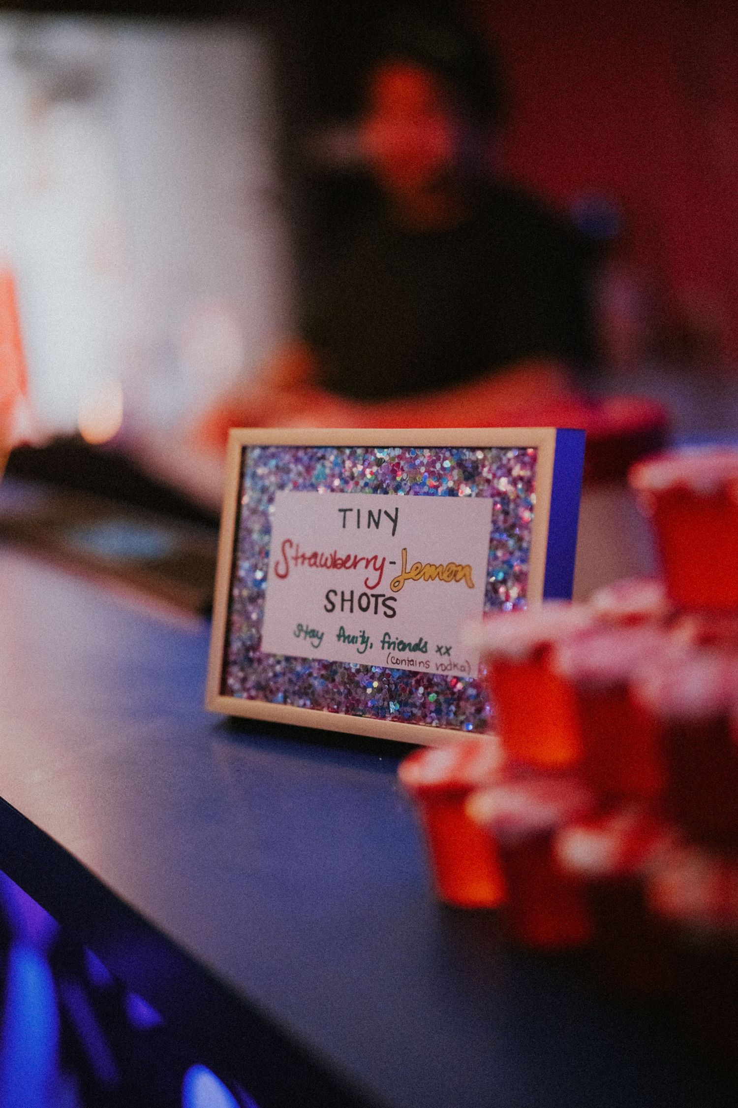 Glitter-framed sign for tiny strawberry lemon shots displayed on a bar next to red plastic cups at a party.