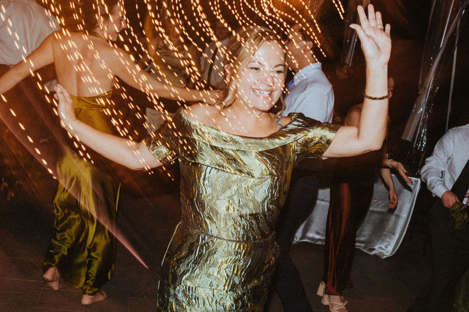 Woman in gold dress dancing joyfully at a wedding reception with bokeh string lights in background.