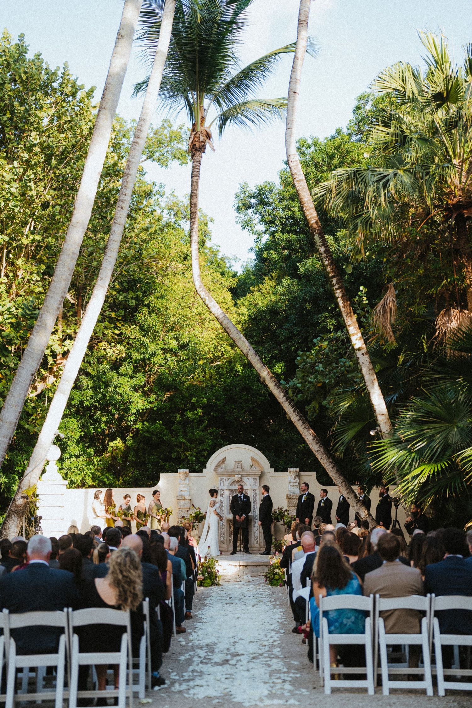 Outdoor wedding ceremony with guests seated in white chairs along a petal-lined aisle beneath tall palm trees.