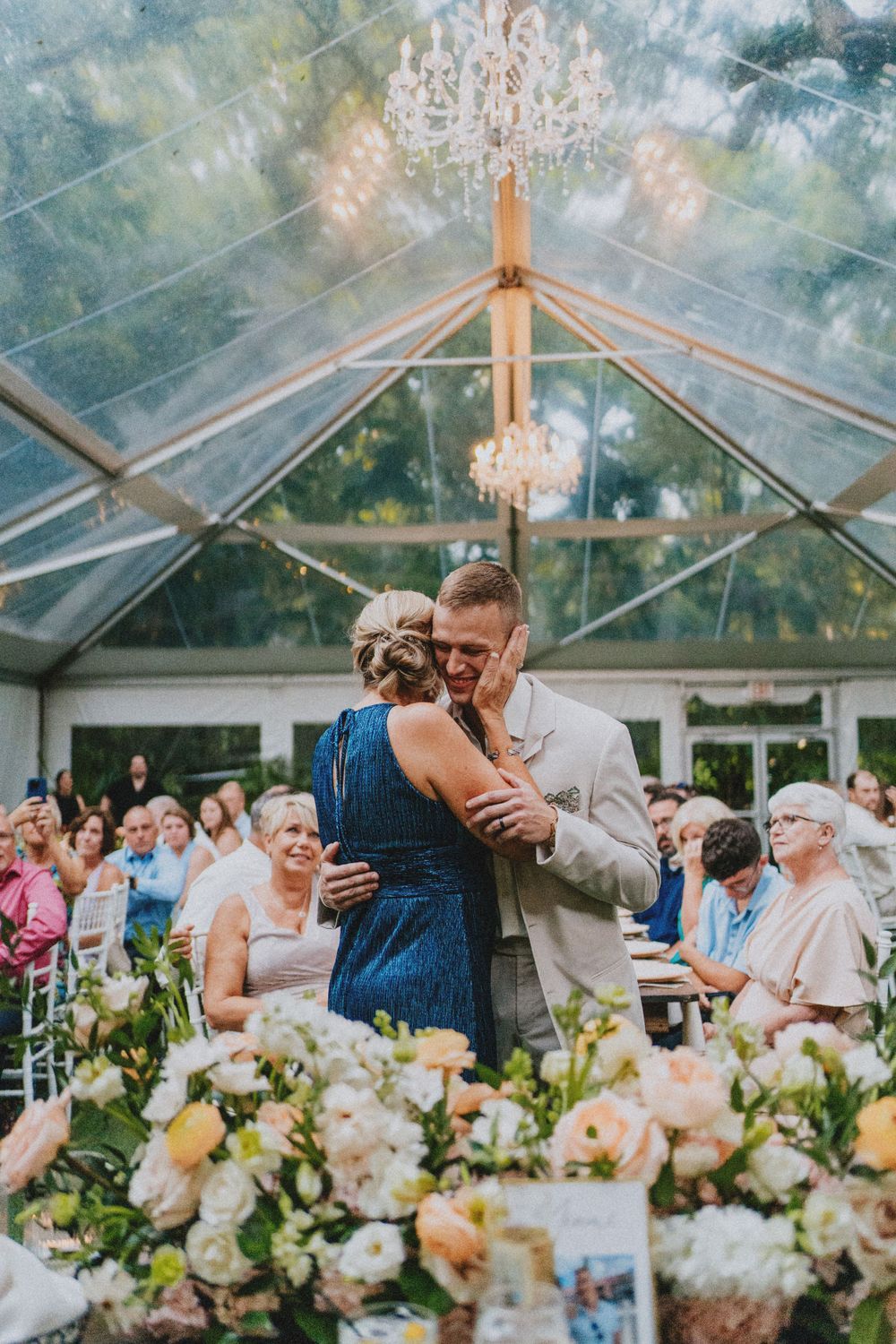 Wedding reception in a glass-roofed venue with elegant floral centerpieces and chandeliers.