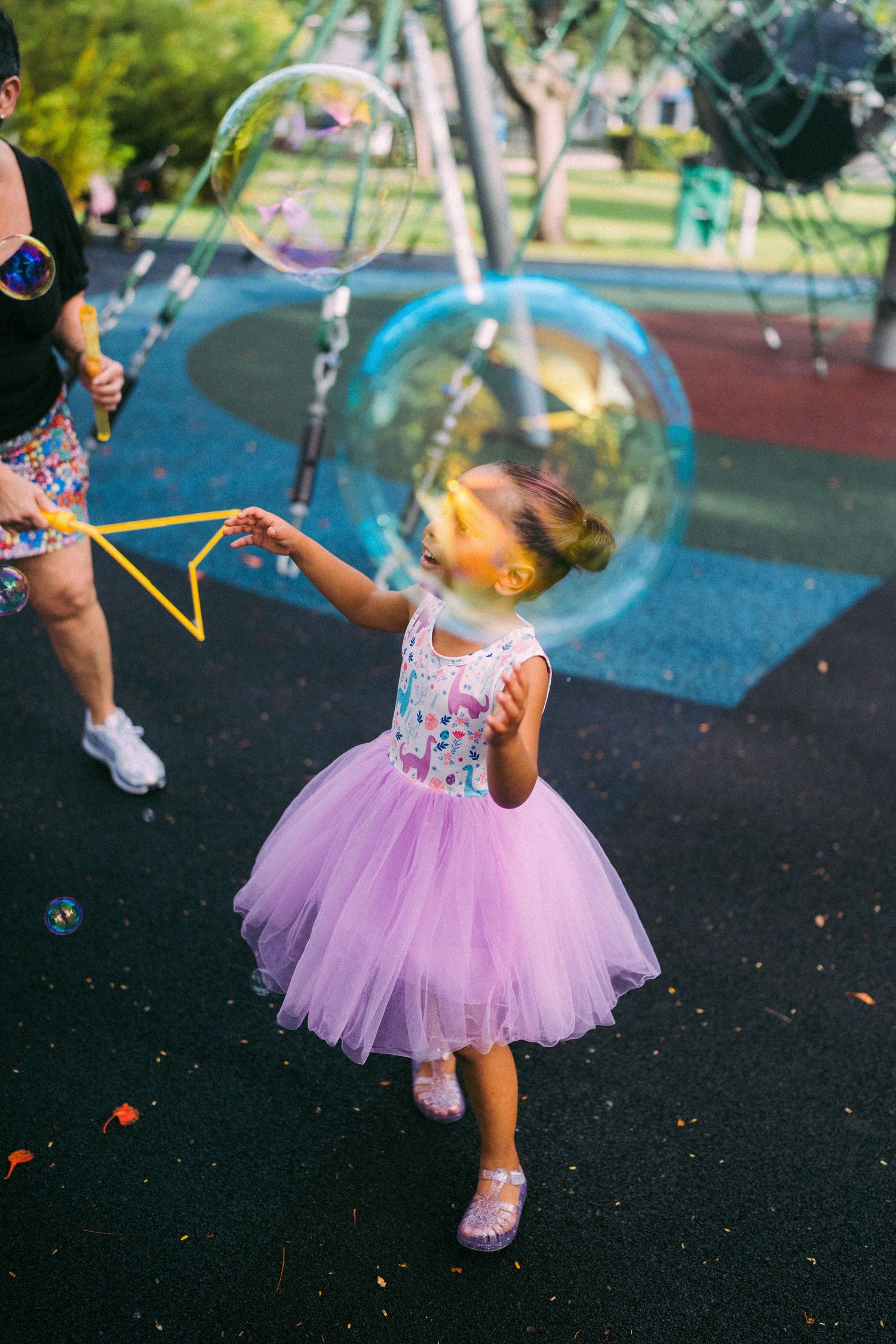 A young girl in a purple tutu dress reaches toward a giant soap bubble at a playground.