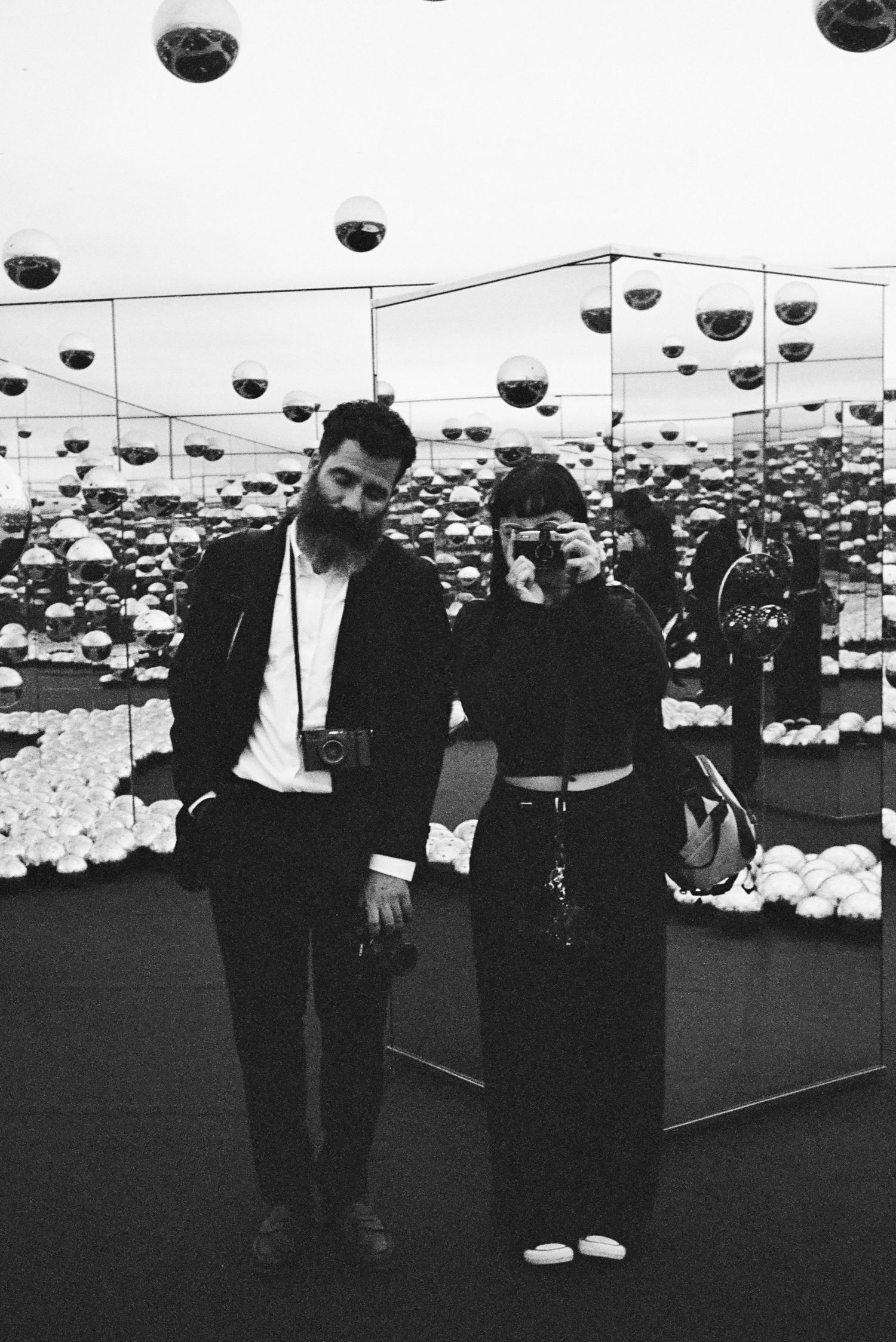 Black and white photo of a bearded man and woman posing in a mirrored room with hanging spherical lights.