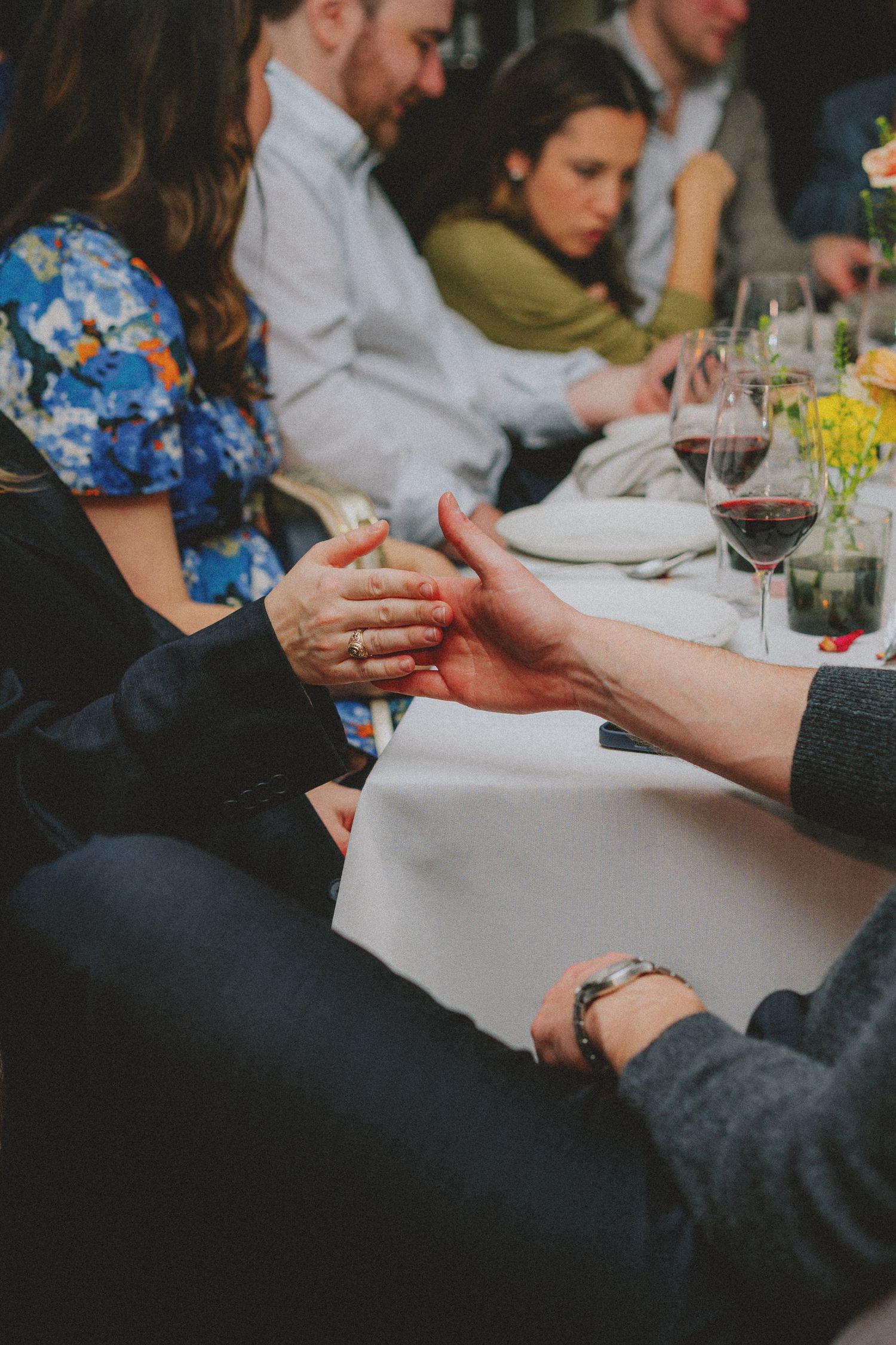 Couple holding hands tenderly at wedding dinner table with wine glasses in background.