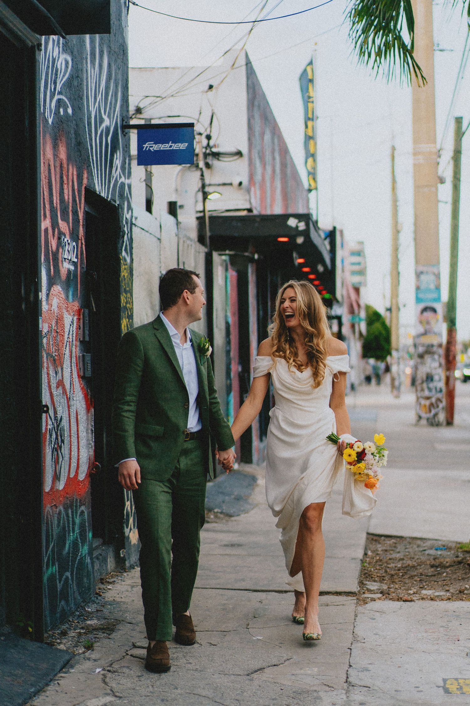 Bride in white dress holding yellow bouquet walks with groom in green suit on graffiti-lined sidewalk.