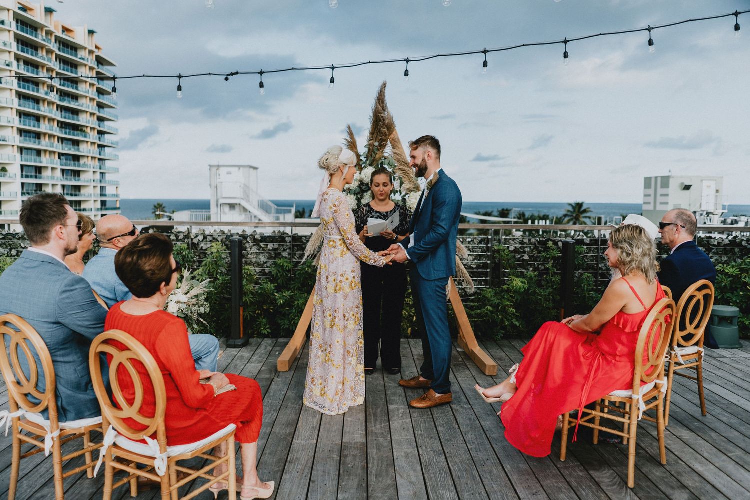Rooftop wedding ceremony with couple exchanging vows, guests in red dresses, string lights, and ocean view backdrop.