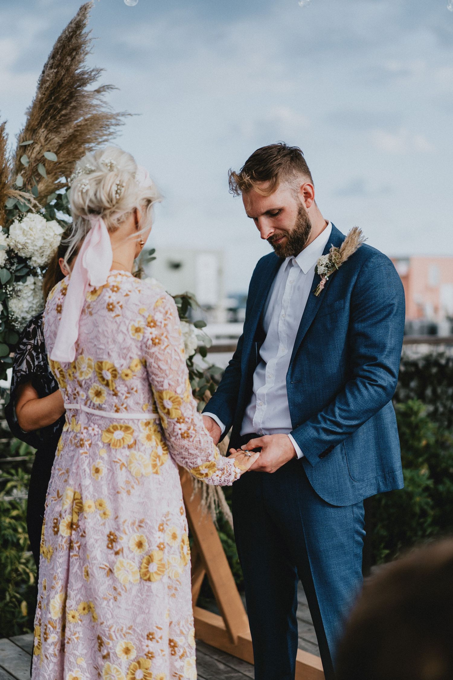 Bride in floral dress and groom in blue suit exchange rings at an outdoor rooftop wedding ceremony.