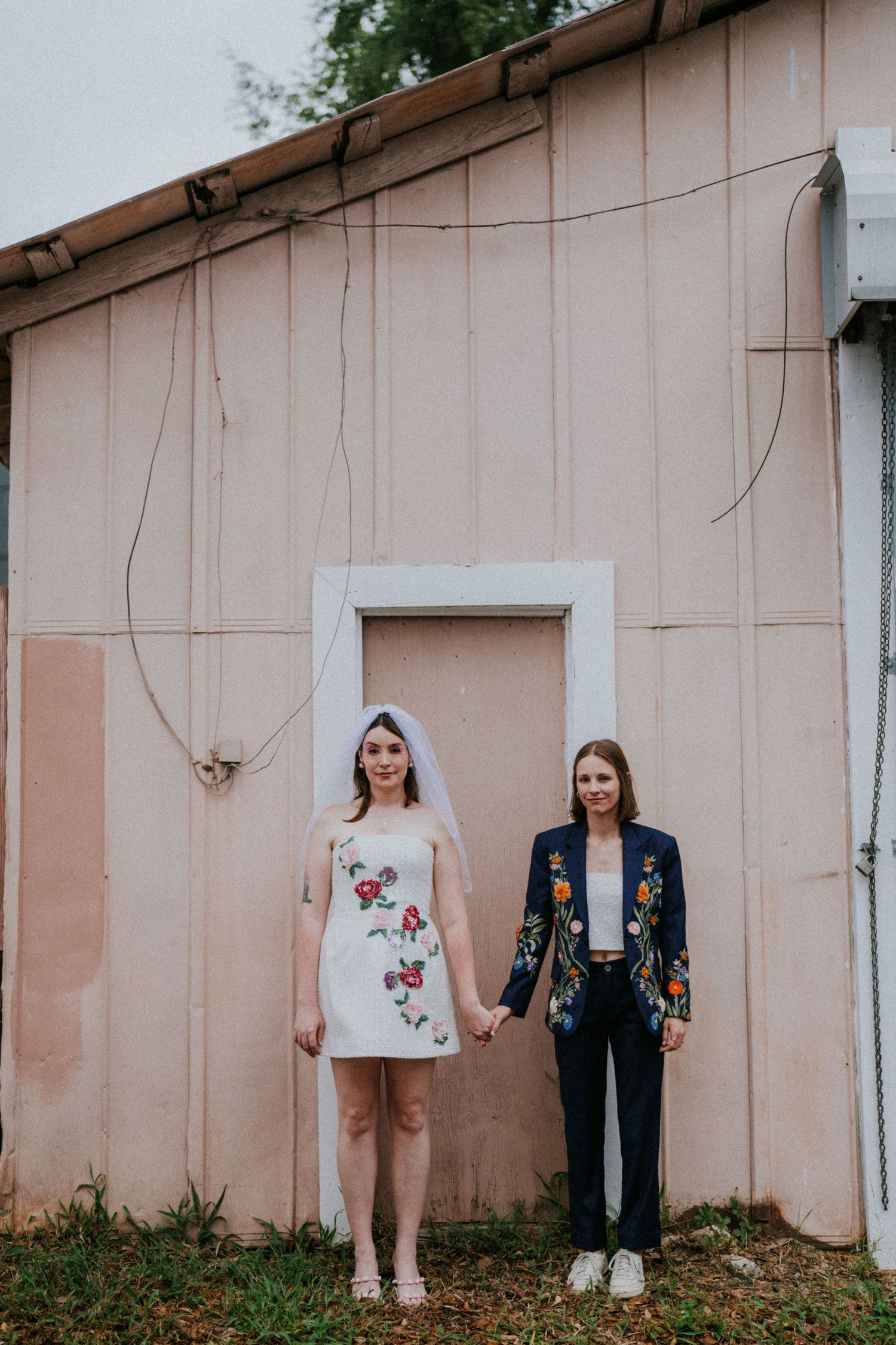 Two brides hold hands outside a pink building, one in a floral mini dress with veil, one in a navy floral suit.