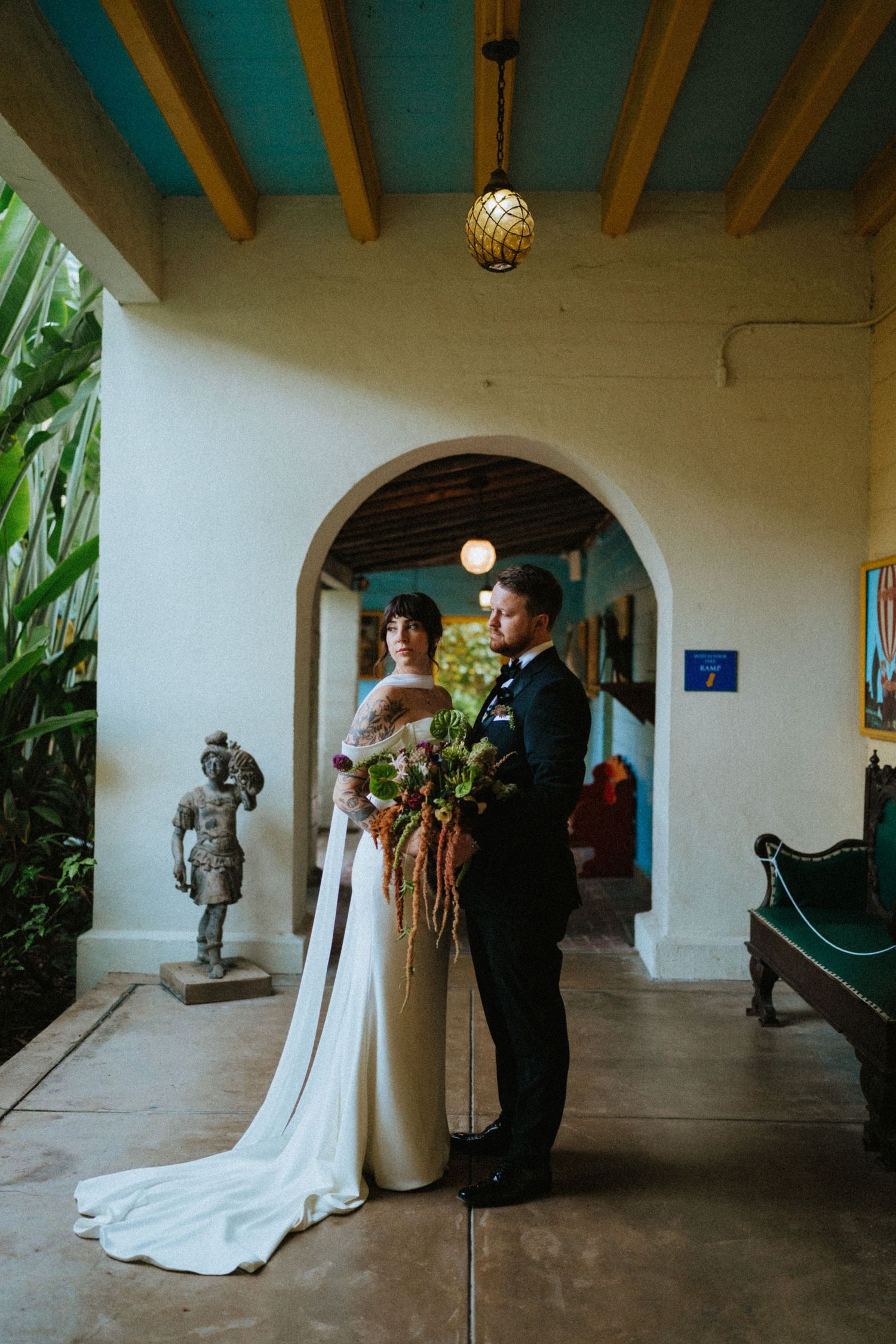 Bride and groom pose in elegant archway, she holds a lush bouquet with long stems, wearing a white gown with train.