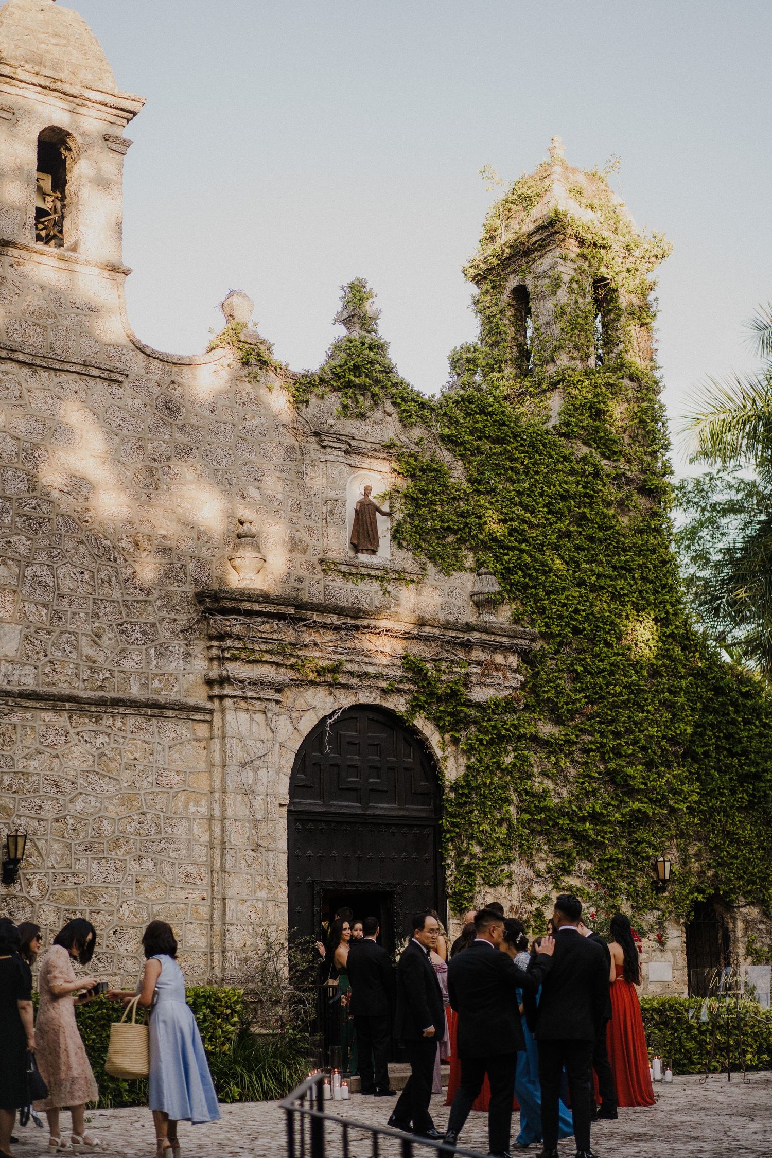 Guests gather outside a historic ivy-covered stone church at golden hour during an elegant outdoor wedding ceremony.