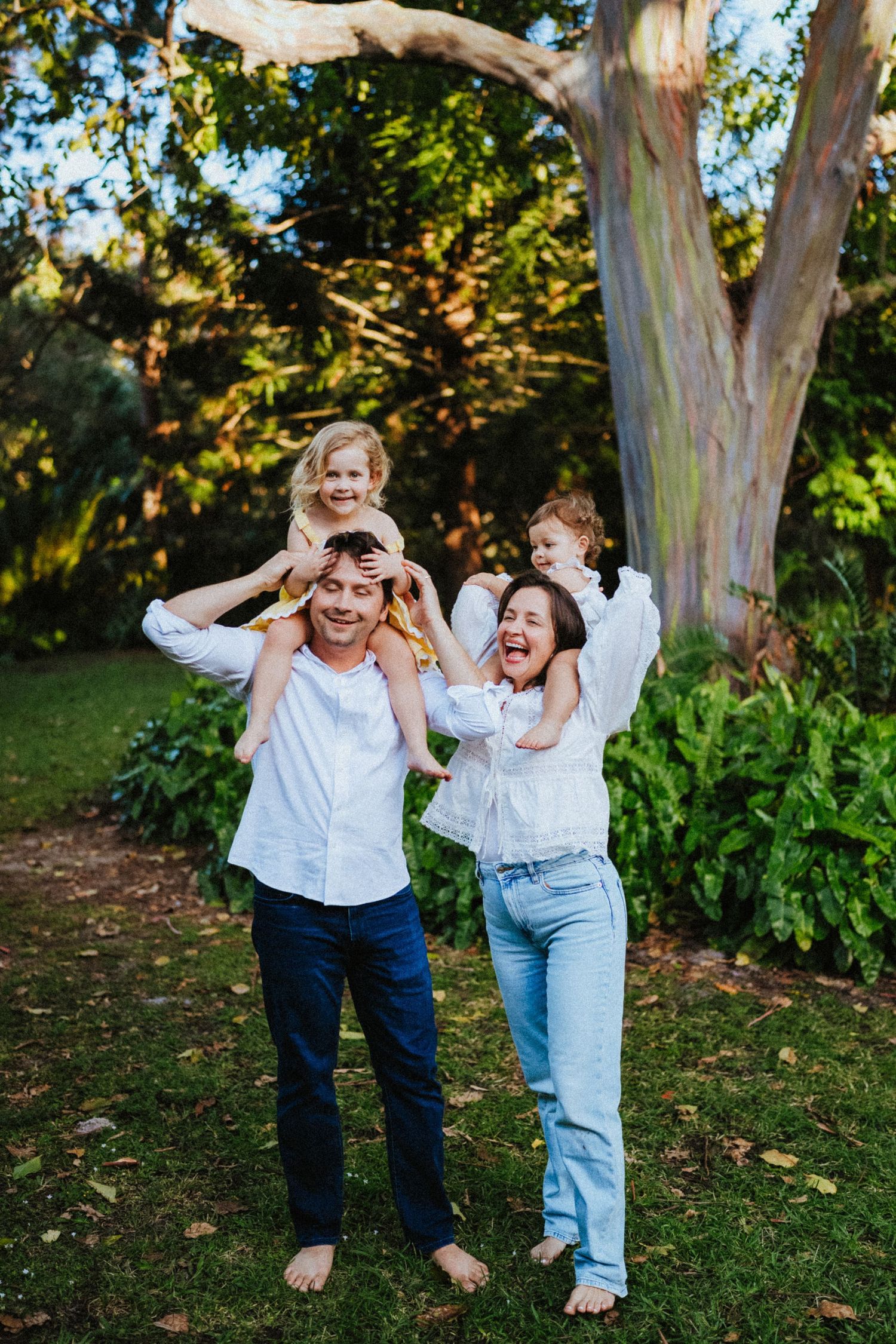 Happy family of four in white shirts laughing outdoors, two children on parents shoulders in lush green garden.