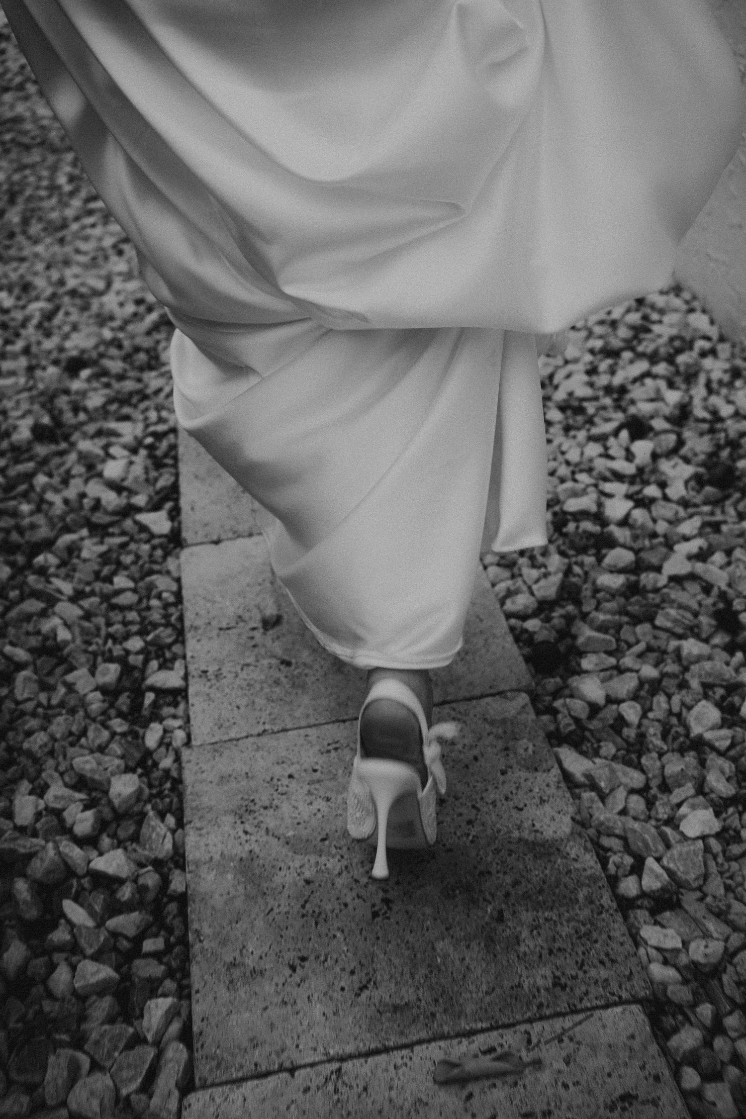 Black and white photo of a bride walking in heels on a stone path, her white gown trailing elegantly behind her.