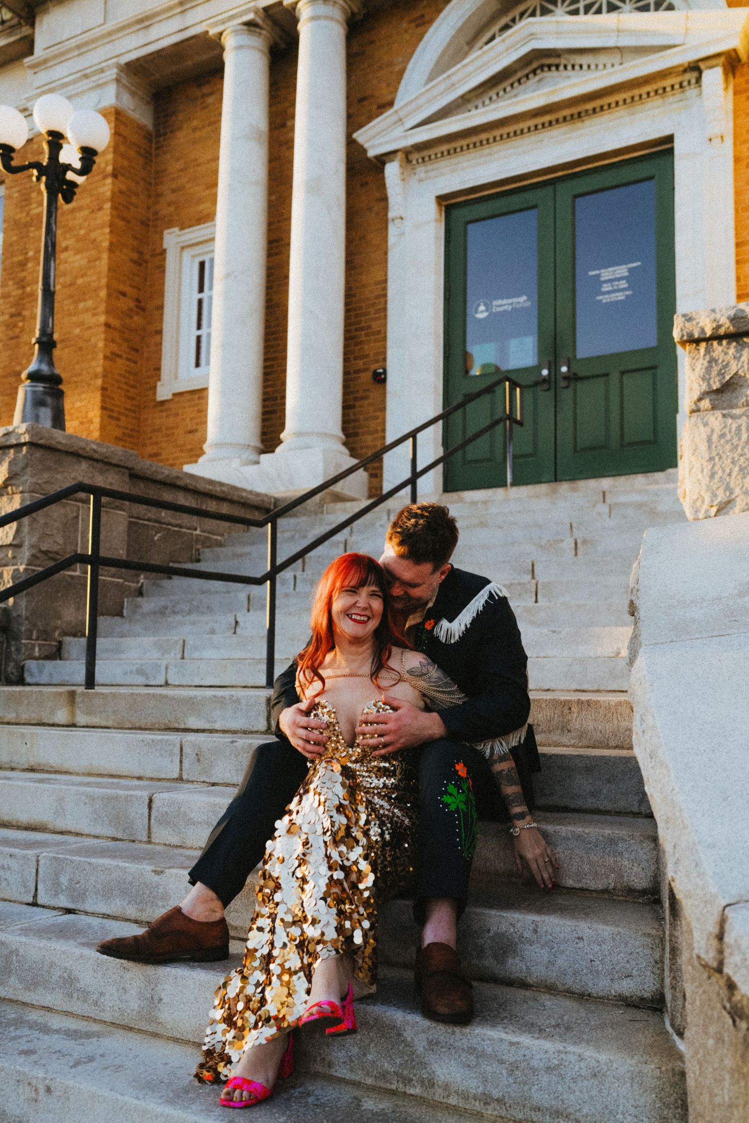 Couple sitting on marble steps of historic building, woman in gold sequin dress with red hair, man embracing her from behind.
