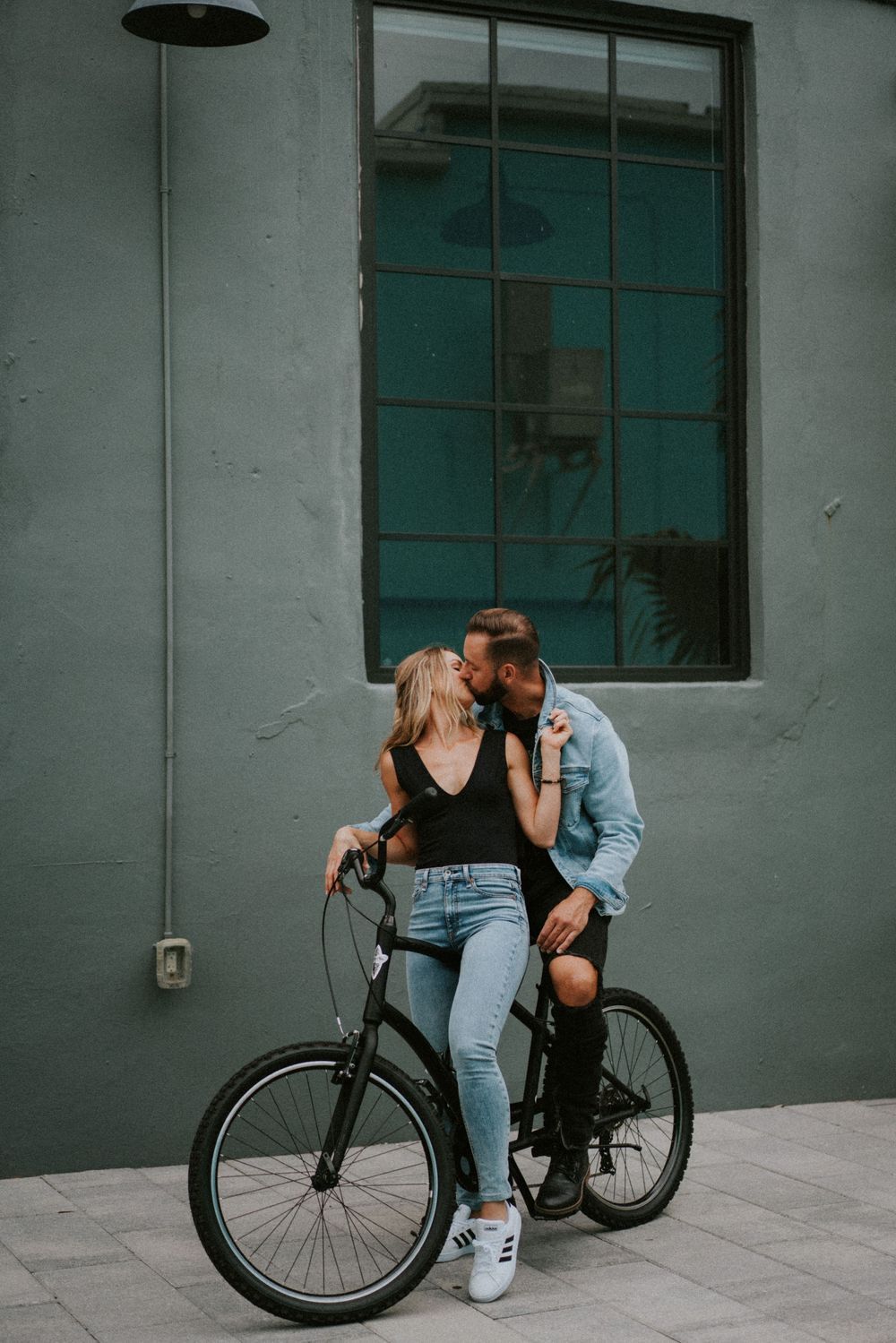 A couple shares an intimate moment while leaning against a bicycle near a gray building.