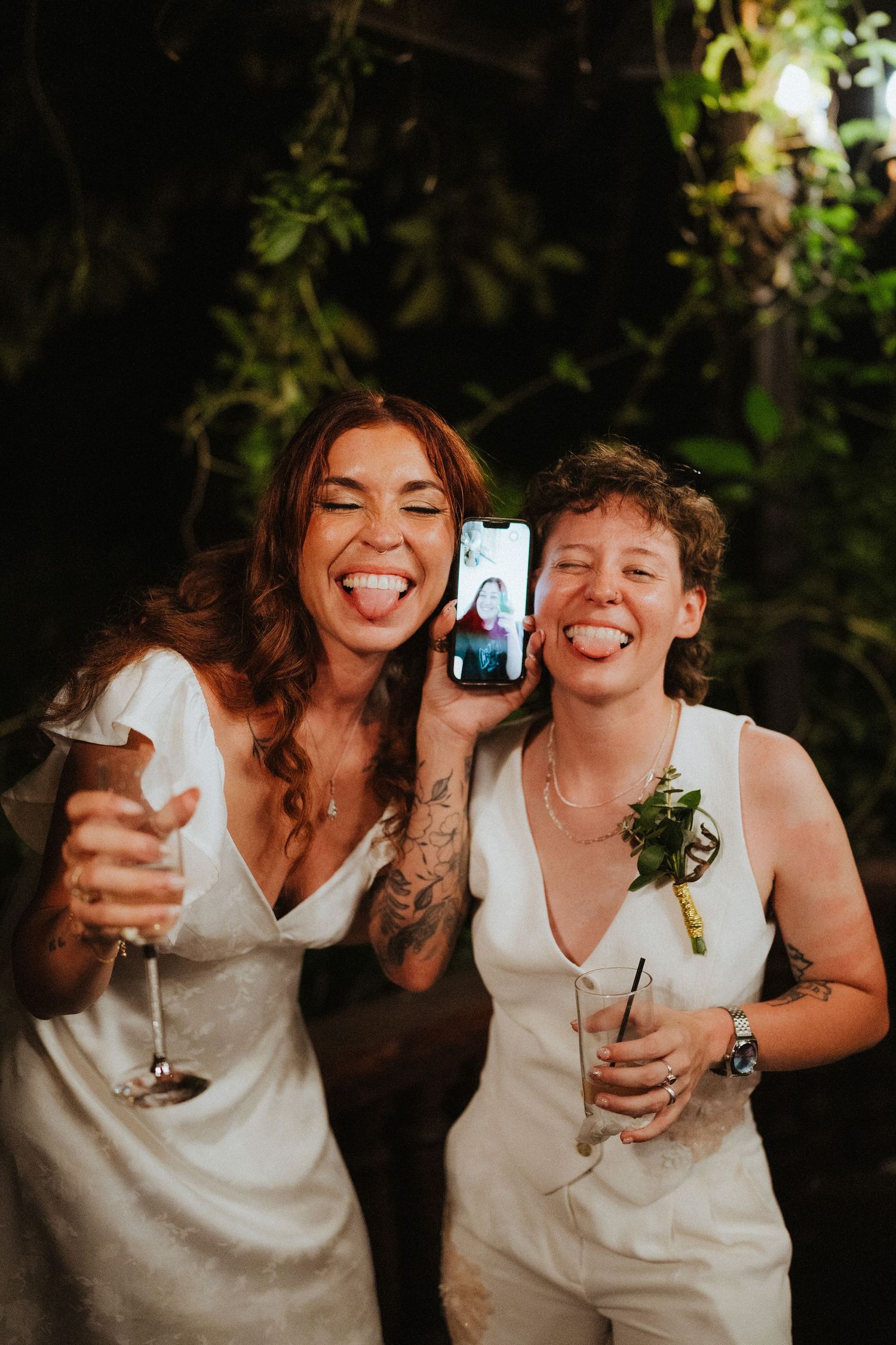 Two brides in white dresses laugh while holding drinks and a phone showing a video call at their wedding reception.