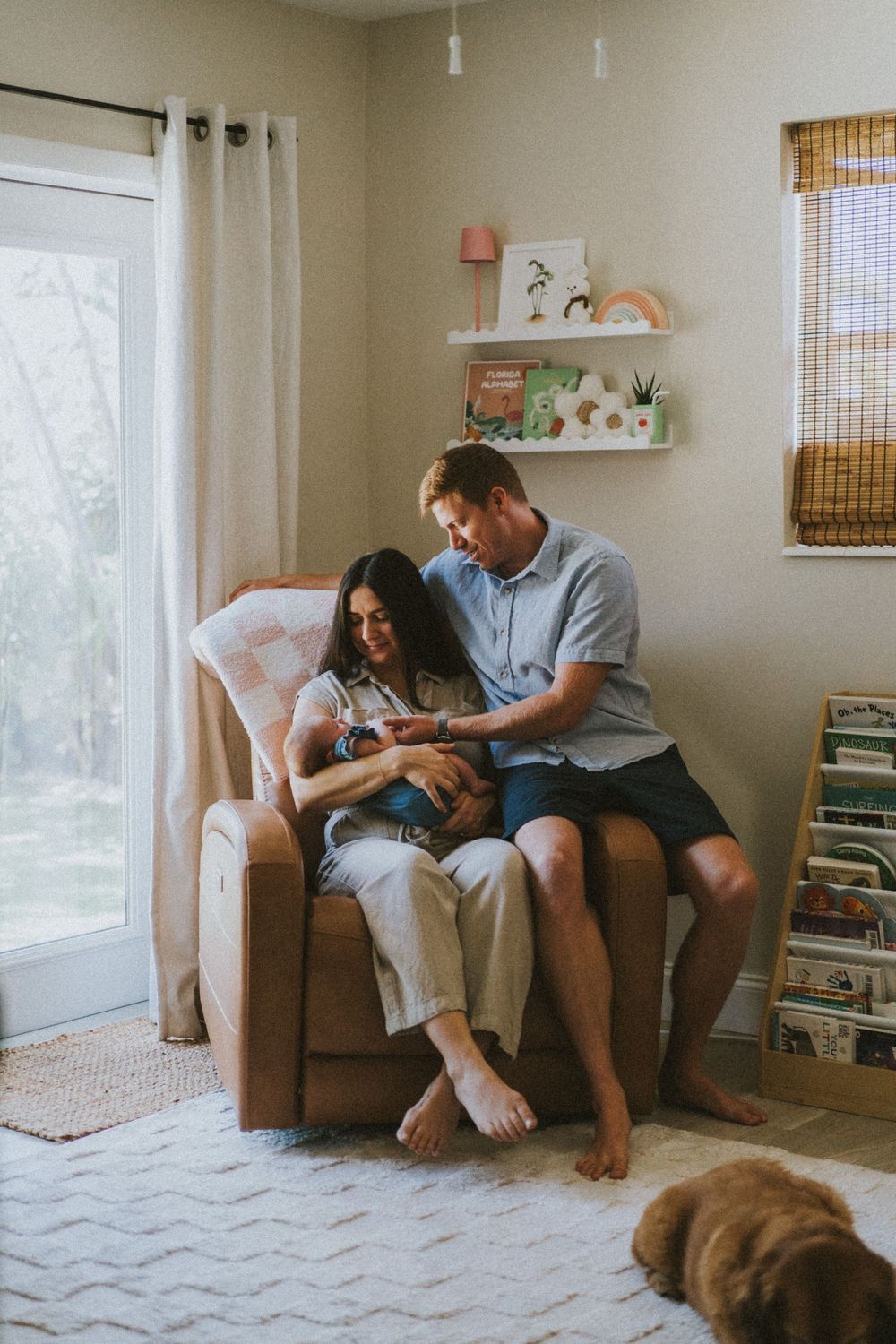 A cozy living room scene with people sitting on a brown couch next to a window and bookshelves.