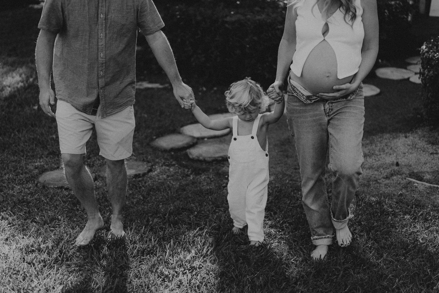 Black and white photo of a toddler holding hands with two adults while walking in a garden setting.