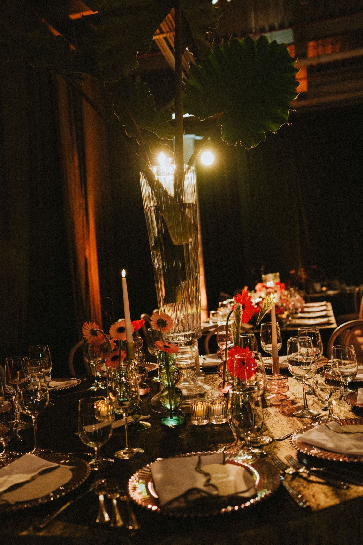 Elegant dark wedding reception table with tropical leaf centerpiece, candles, crystal glasses, and red floral arrangements.
