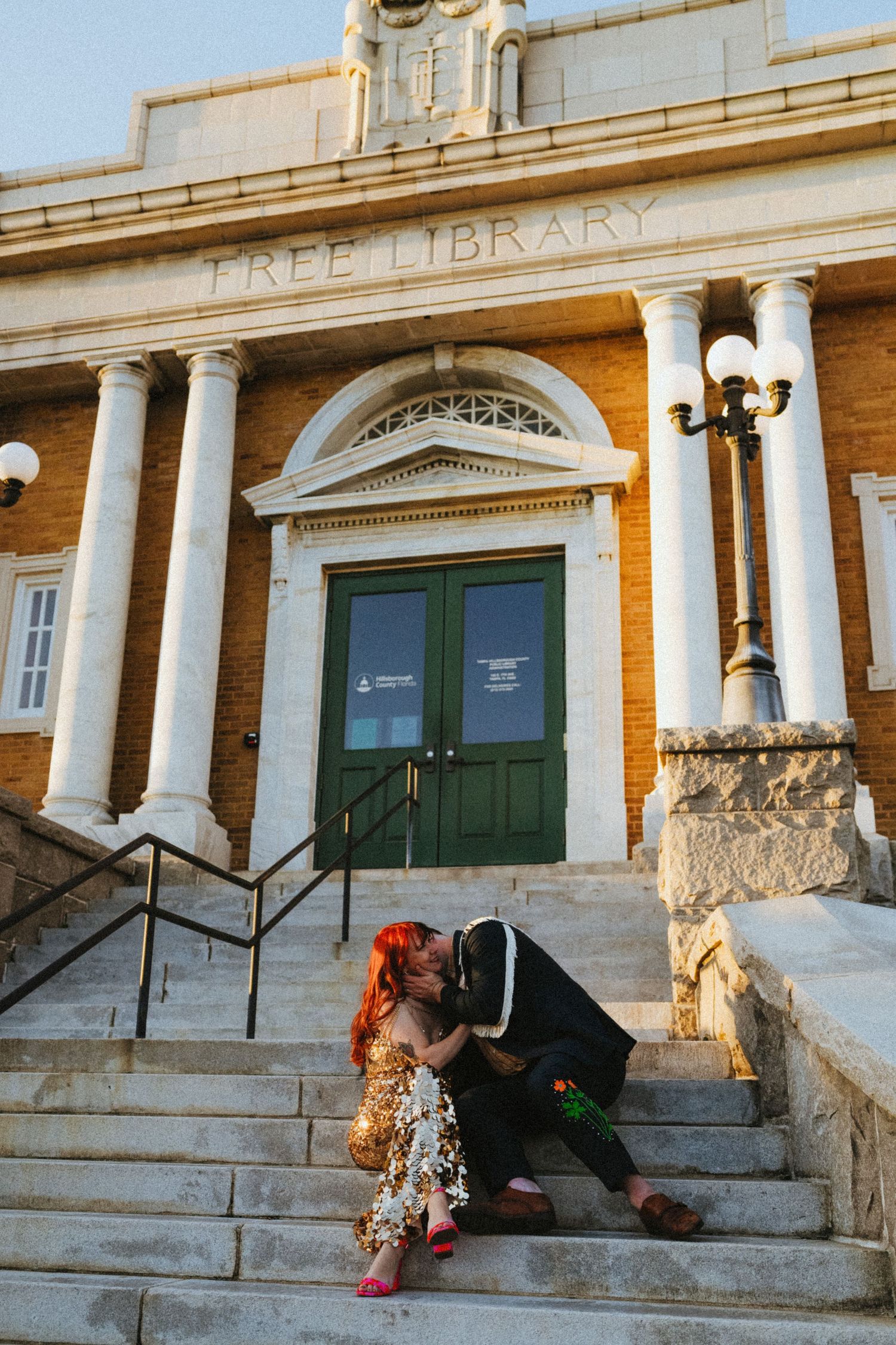 Couple sharing a romantic kiss on the steps of a classic brick library with white columns.