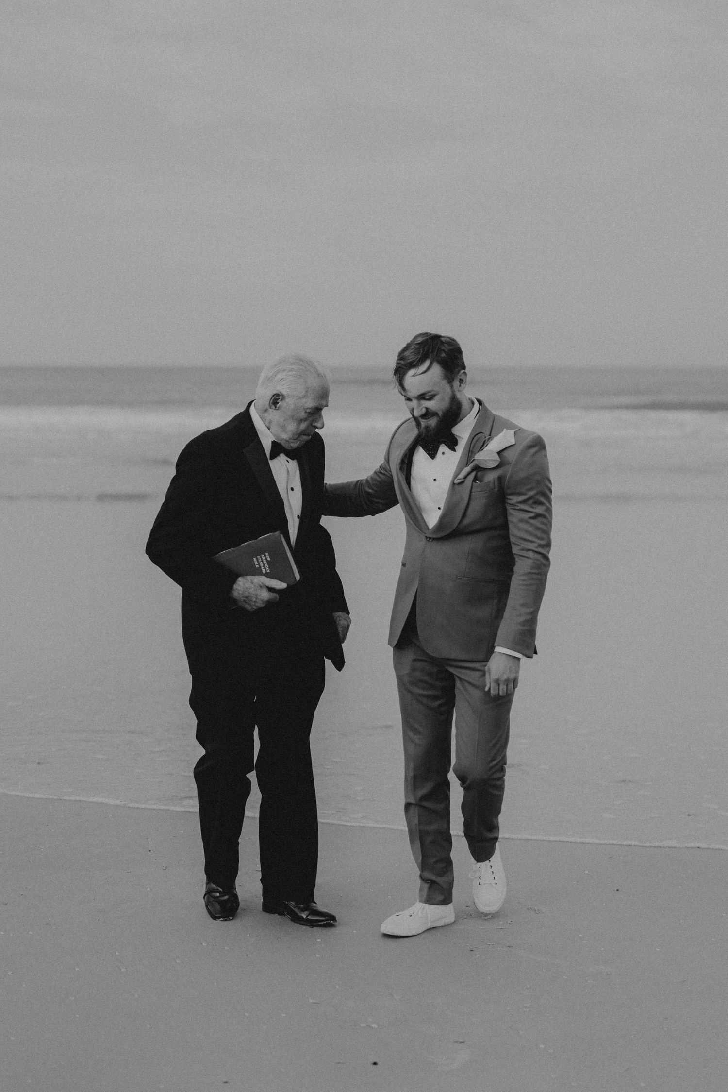Groom in mustard suit speaking with elderly officiant on beach in black and white.