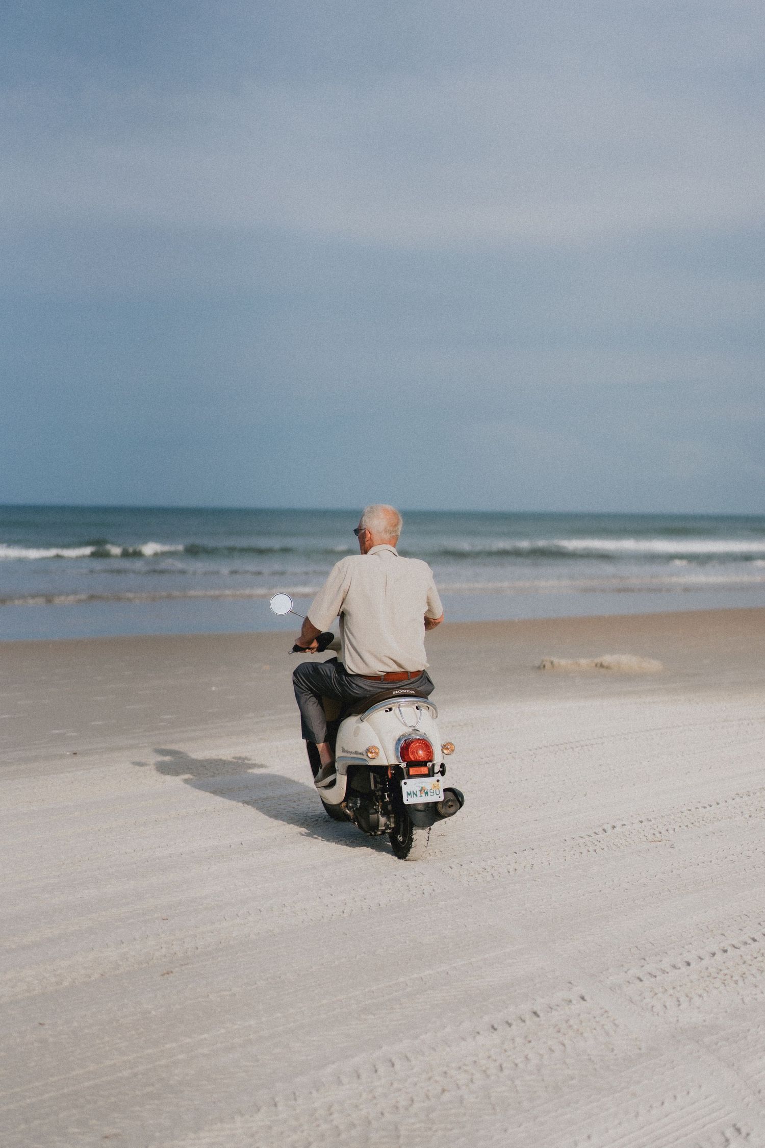 Elderly man riding vintage scooter along sandy beach with ocean waves behind.