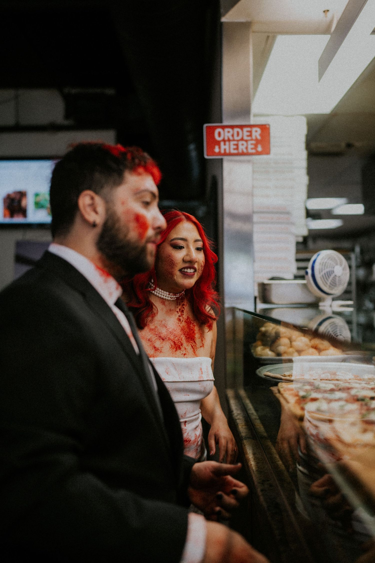 Zombie wedding couple with bloody Halloween makeup ordering pizza at a restaurant counter.