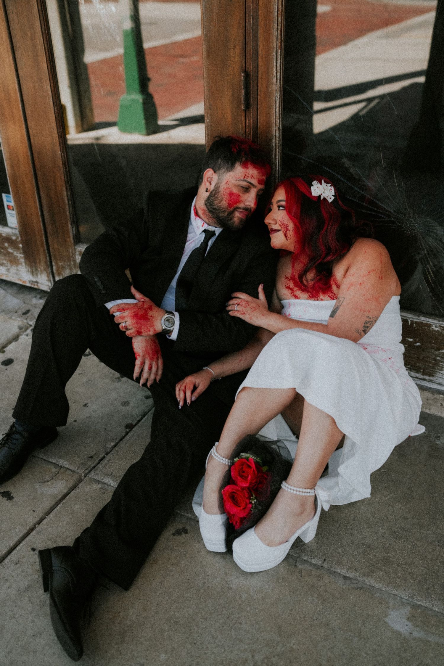 Bride with red hair and groom in black suit sit together on sidewalk, adorned with red roses and paint.