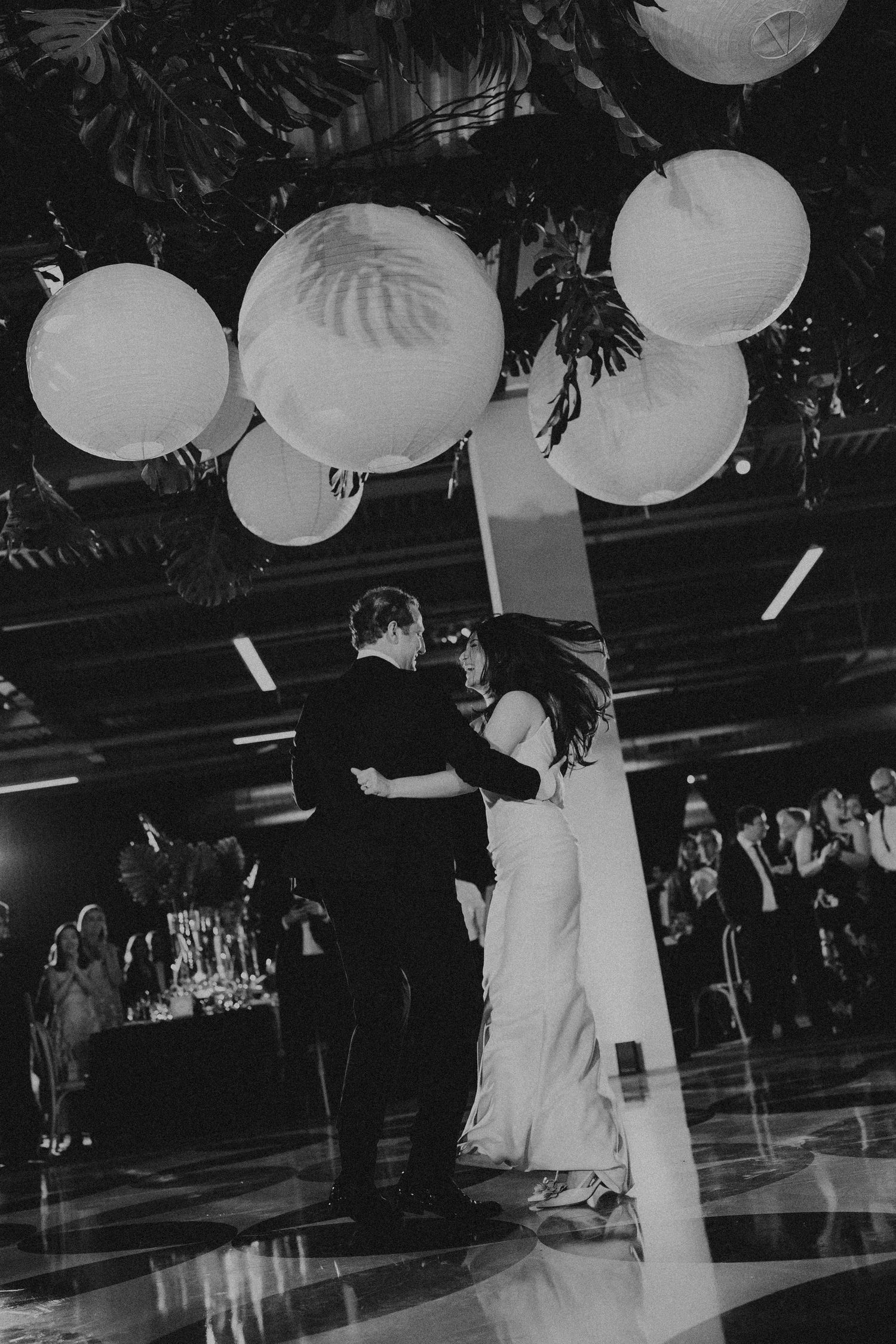 Black and white photo of a couple sharing their first dance under glowing paper lanterns at an elegant wedding reception.