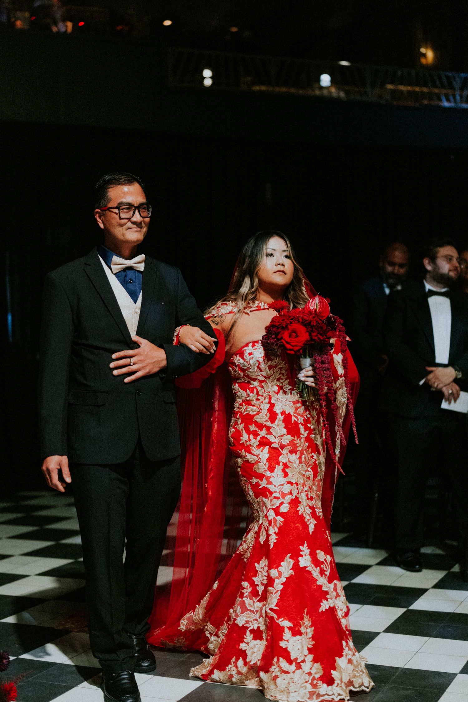 Bride in red and gold lace gown with cape holding red roses walks with escort on checkered floor.